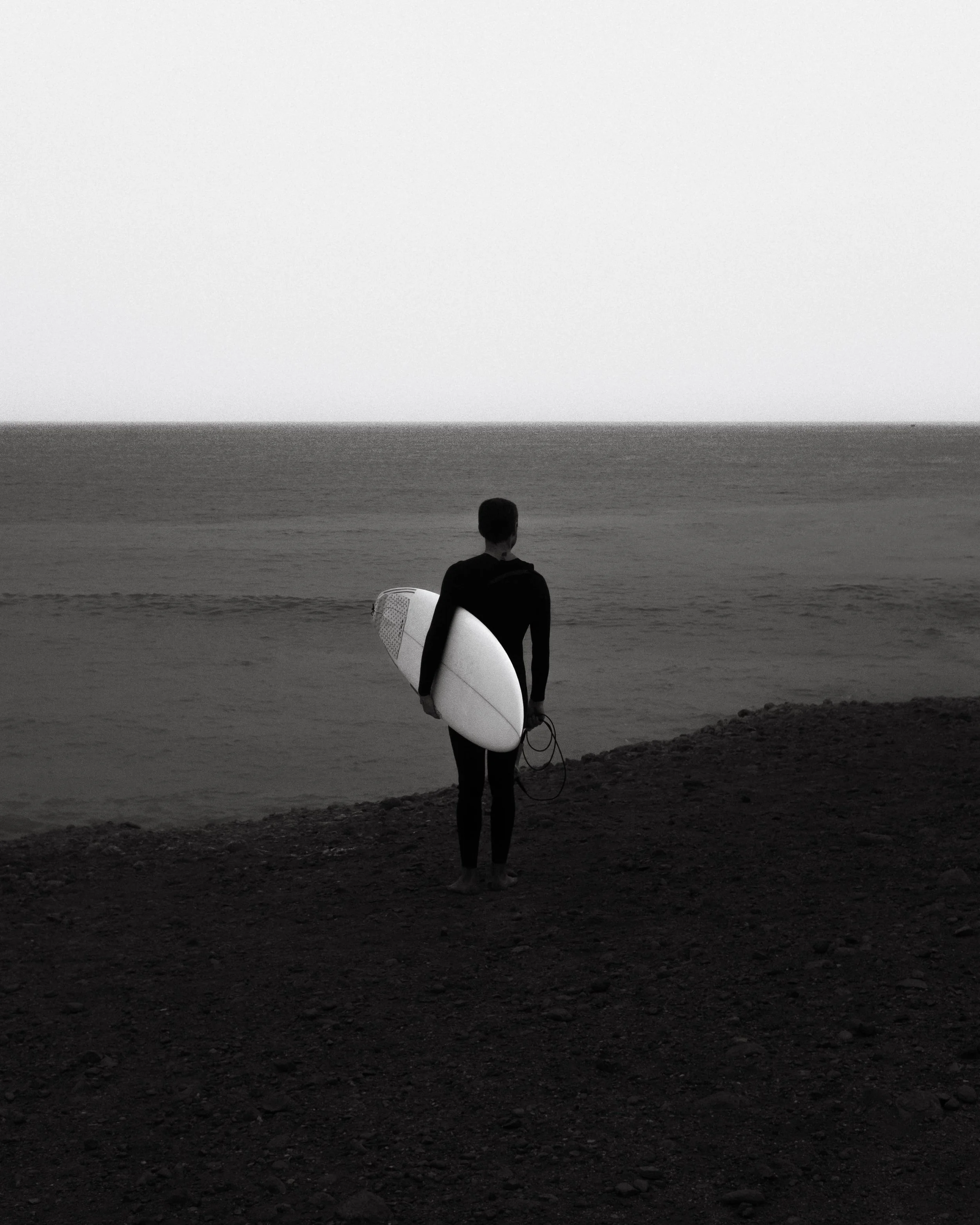 A person standing on a rocky beach holding a surfboard and a leash, looking out at the ocean at dusk or dawn.