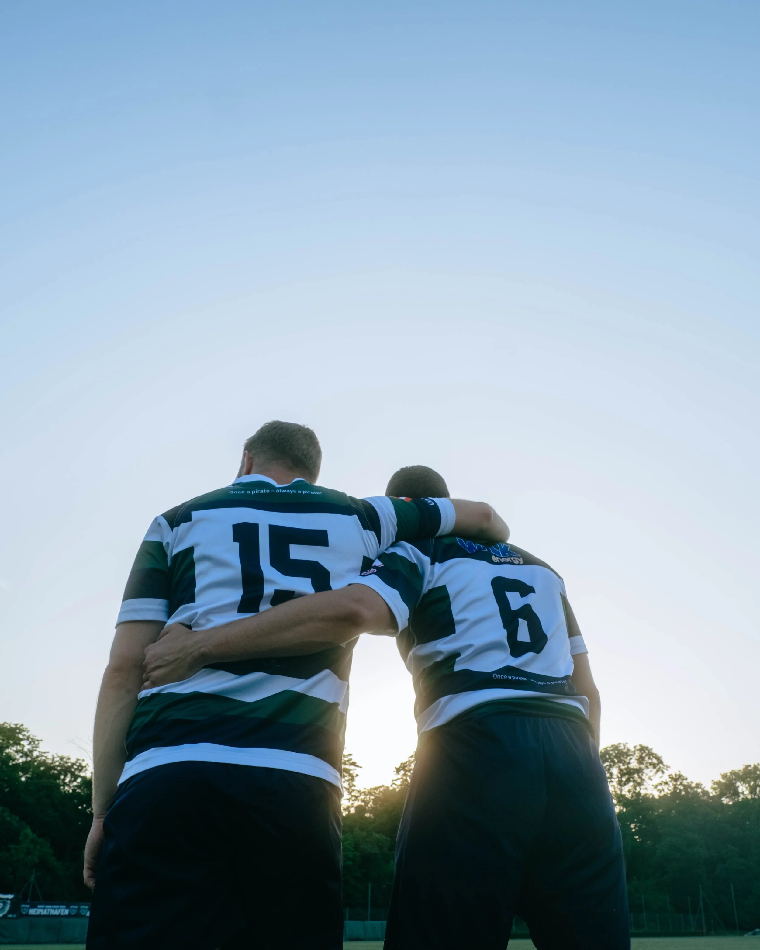 Two soccer players in black and white striped jerseys are standing close together on a field, with one player's arm around the other's shoulder, during sunset.