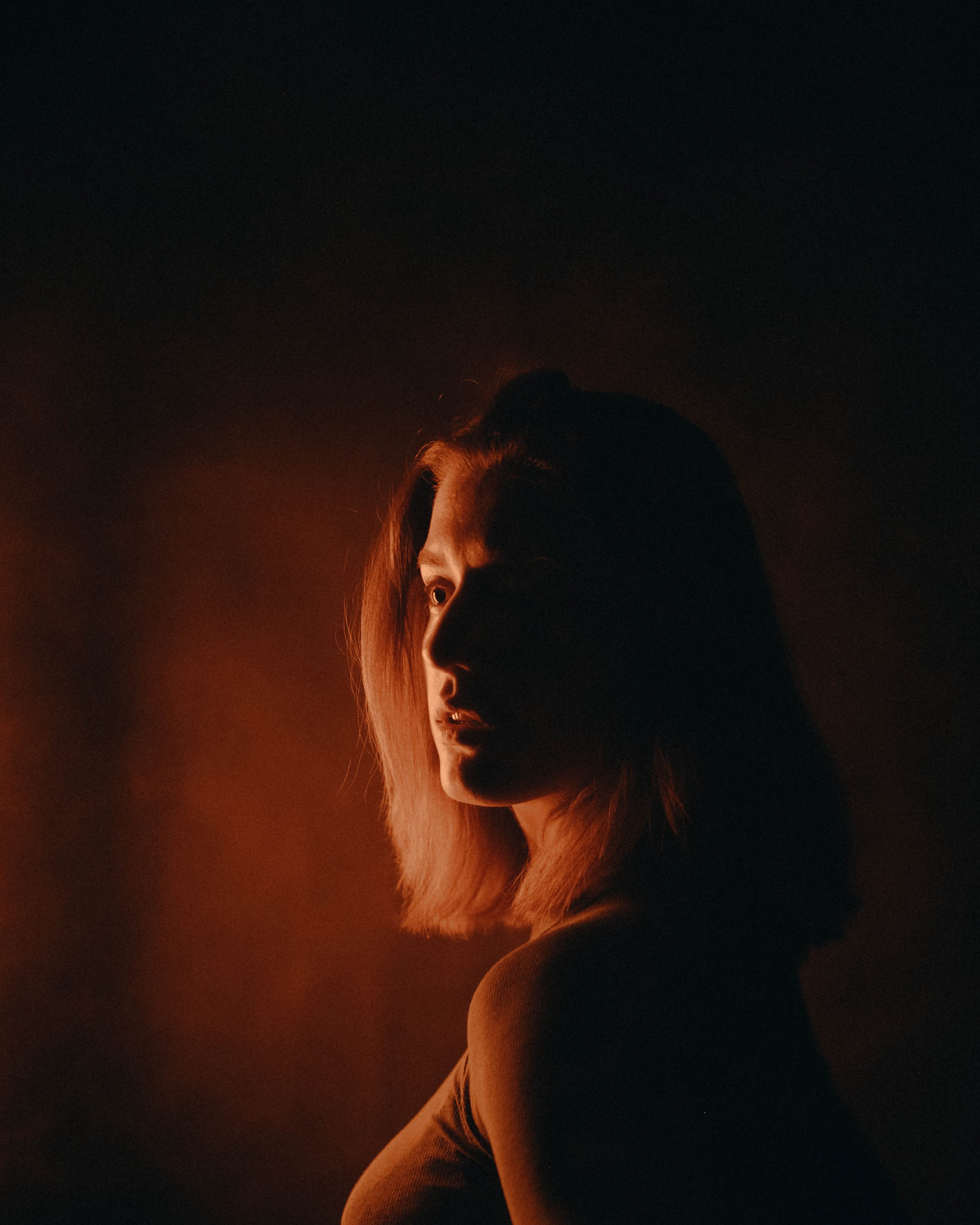 A woman with shoulder-length hair looking to the side, illuminated by warm light from the left, with a dark background.