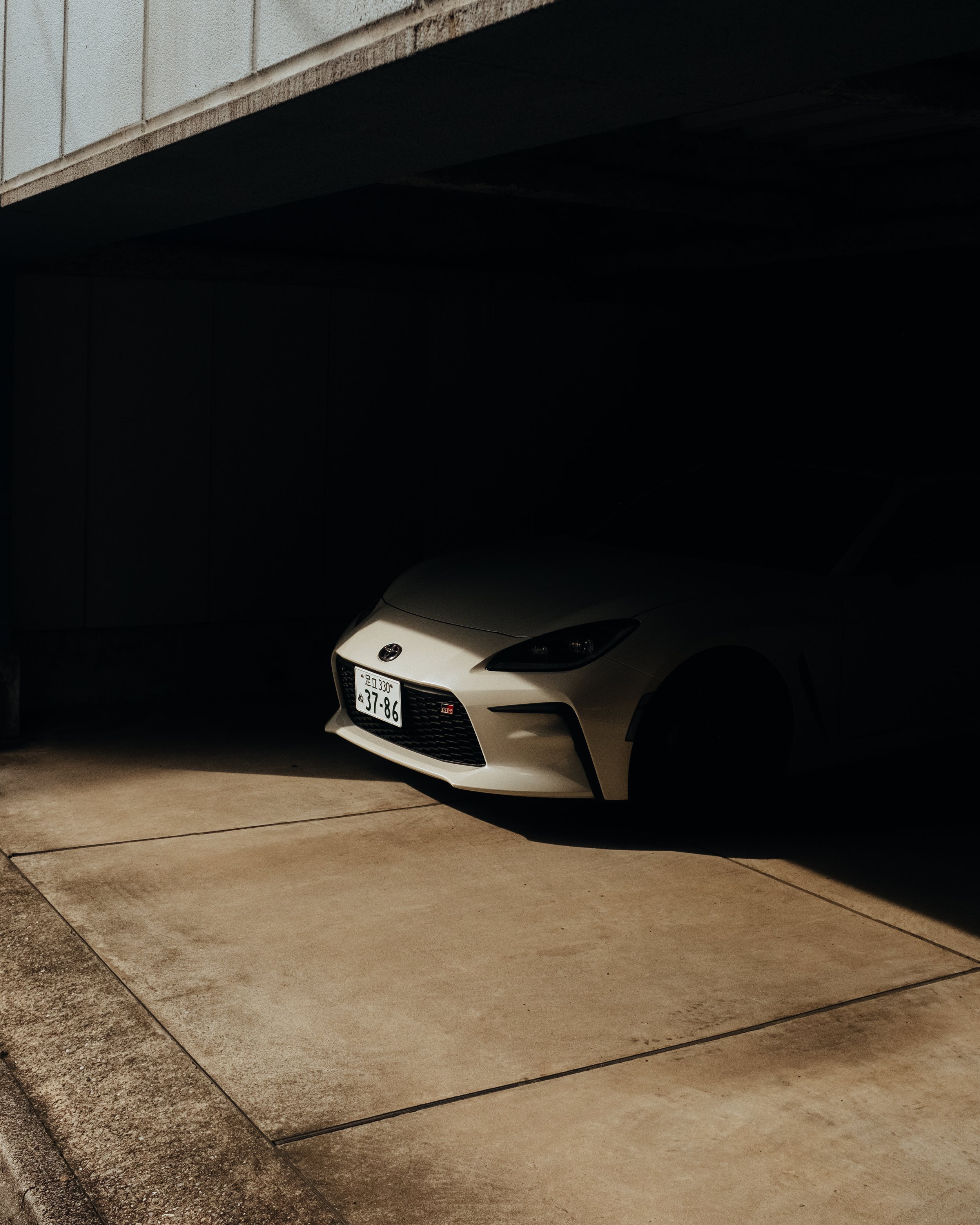 A white sports car parked in a dark garage with sunlight illuminating the front part of the car.
