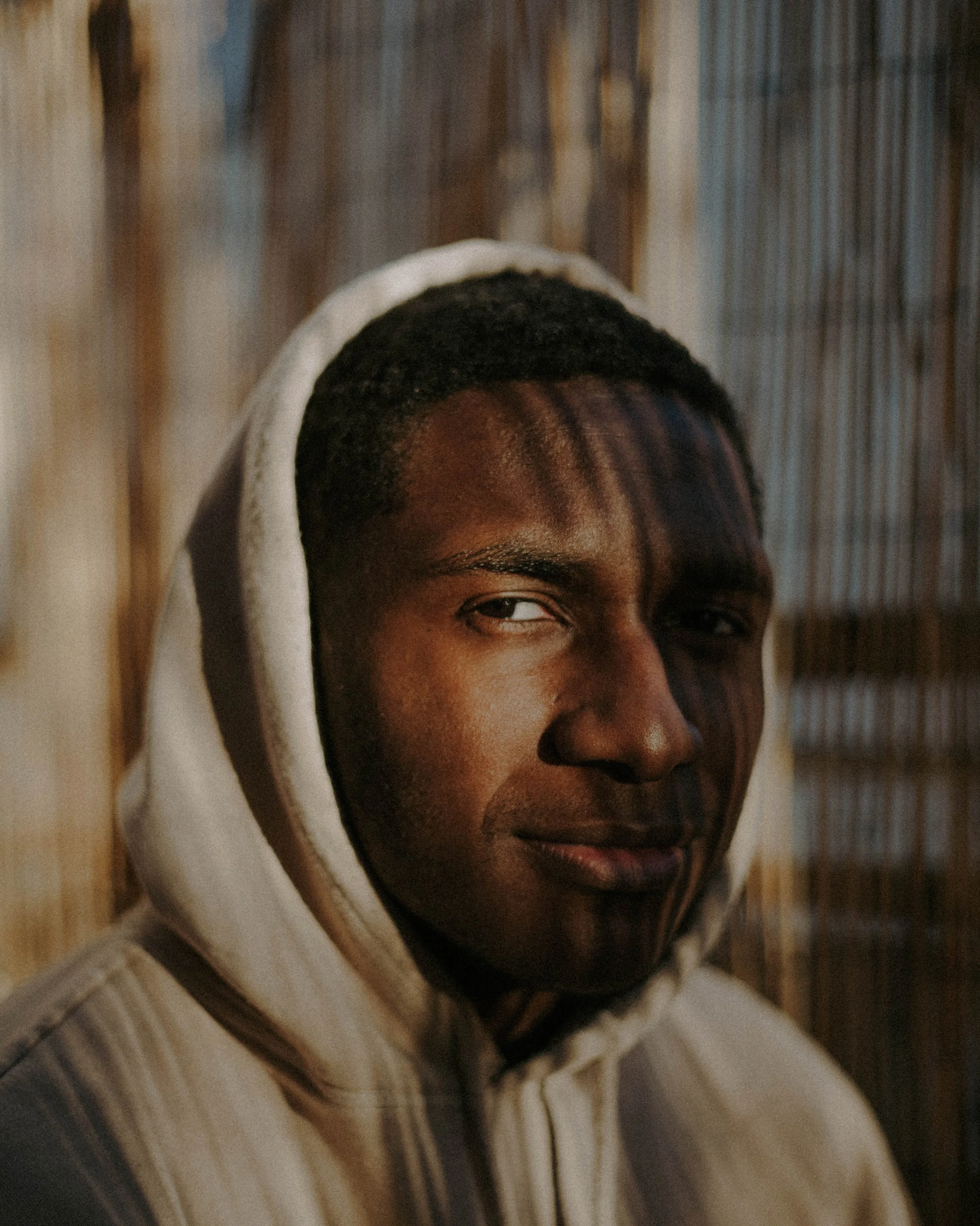 A young man wearing a beige hoodie, looking sideways with shadows cast on his face from a nearby fence or structure.