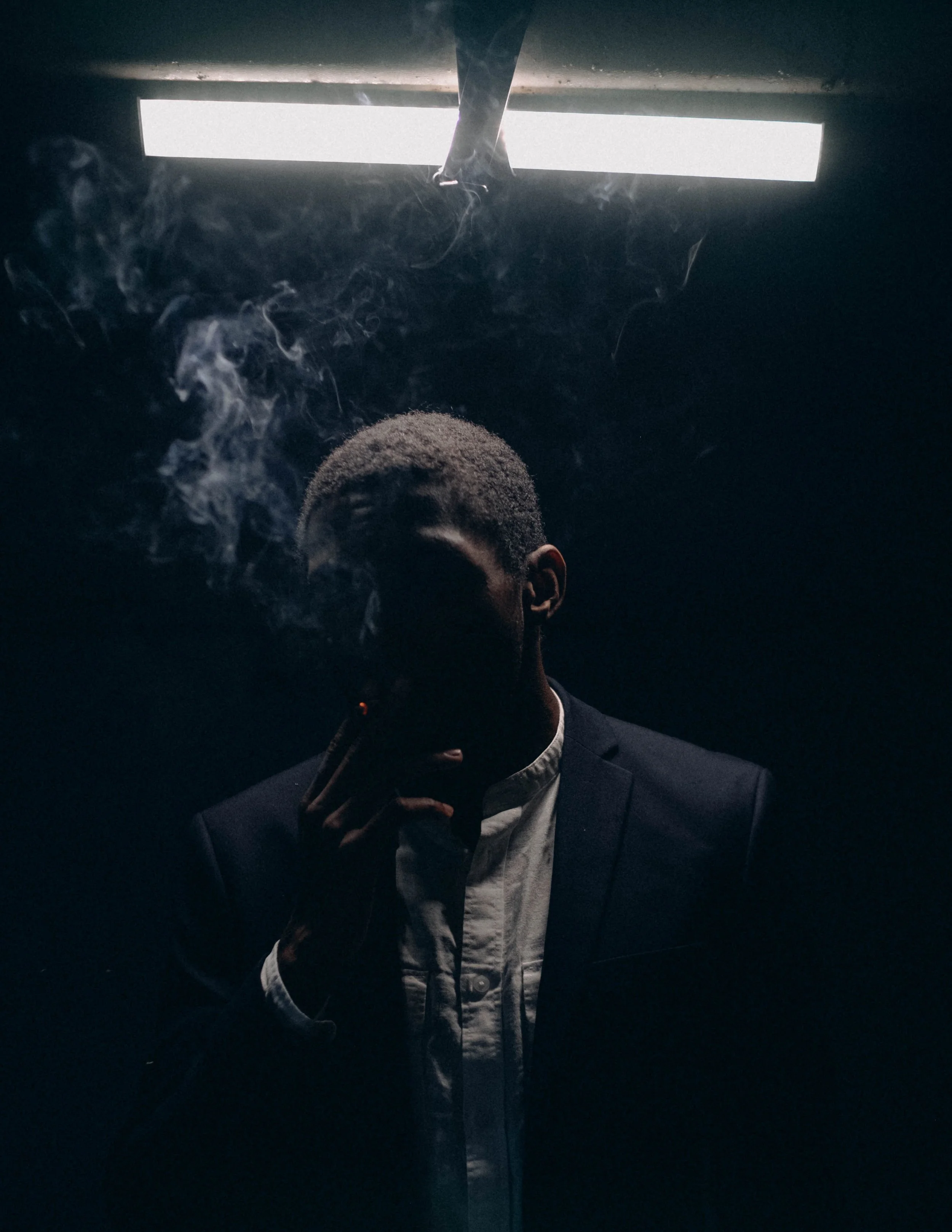 A man in a dark suit is smoking and holding a cigarette. The scene is dimly lit with a bright light source above, creating a moody atmosphere.