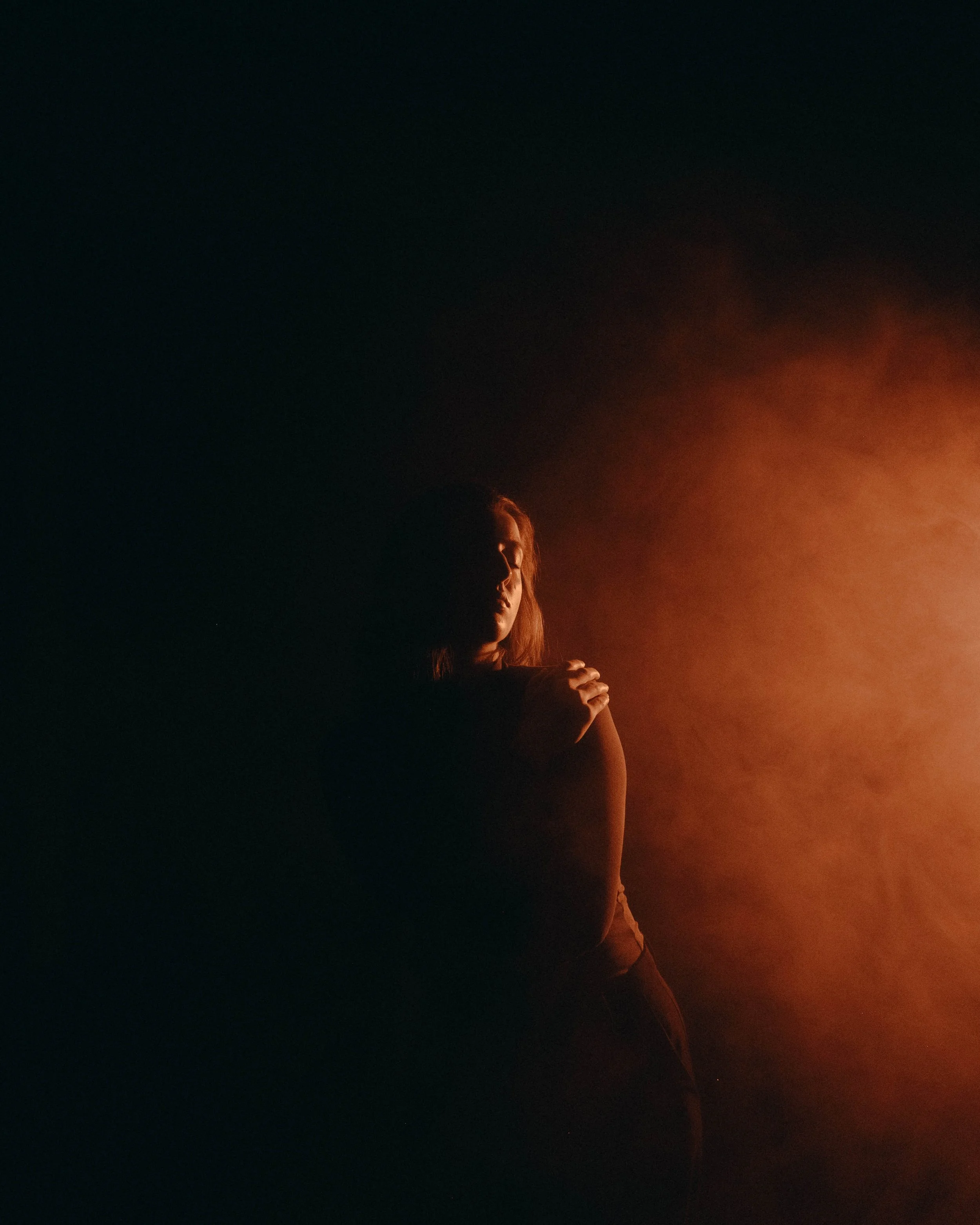 A woman standing with her arms crossed, illuminated by a warm orange light on her shoulder, in a dark setting with a smoky background.
