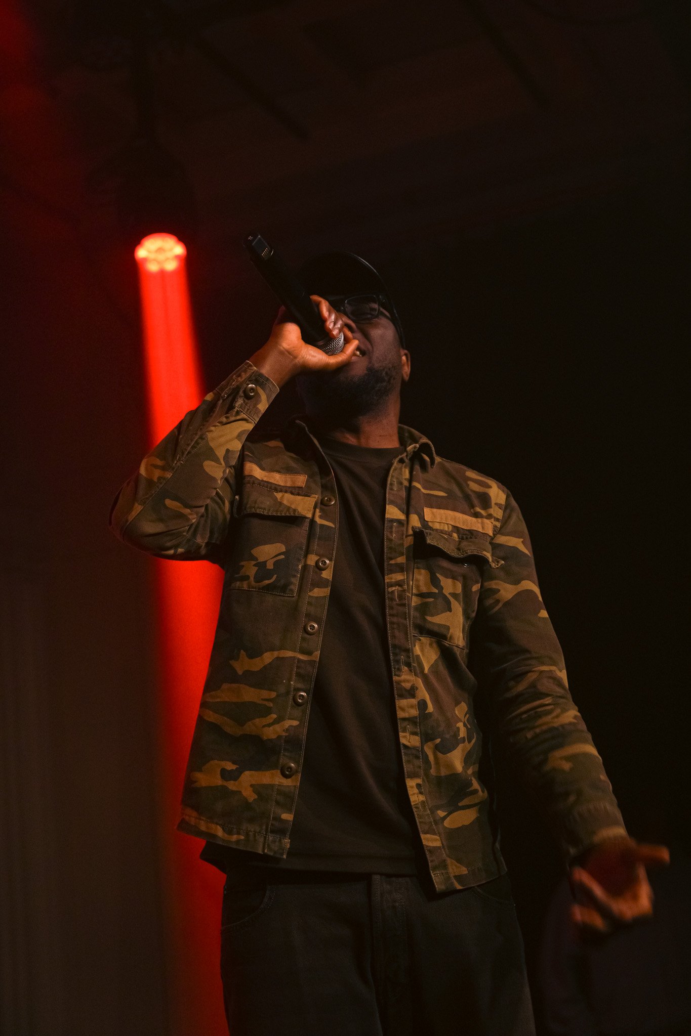 Kendan Adams singing into microphone on stage, wearing camouflage jacket, black shirt, black cap, and sunglasses, with red stage light in background.