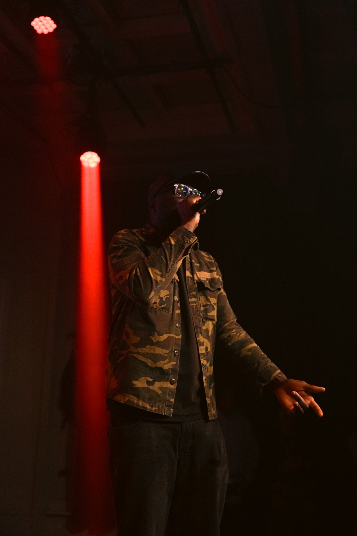 Kendan Adams wearing a camouflage jacket, black shirt, and glasses singing or speaking into a microphone on a dark stage with red lighting.