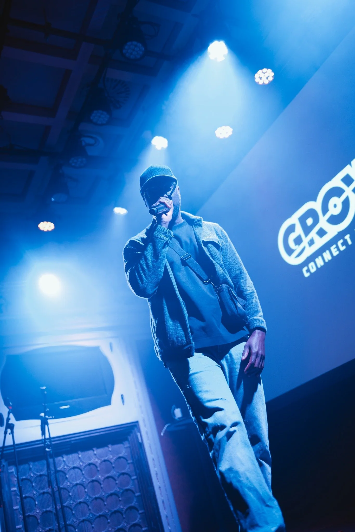 Kendan Adams on stage, wearing sunglasses, a hat, and casual clothing, illuminated by blue stage lights in front of a large screen display.