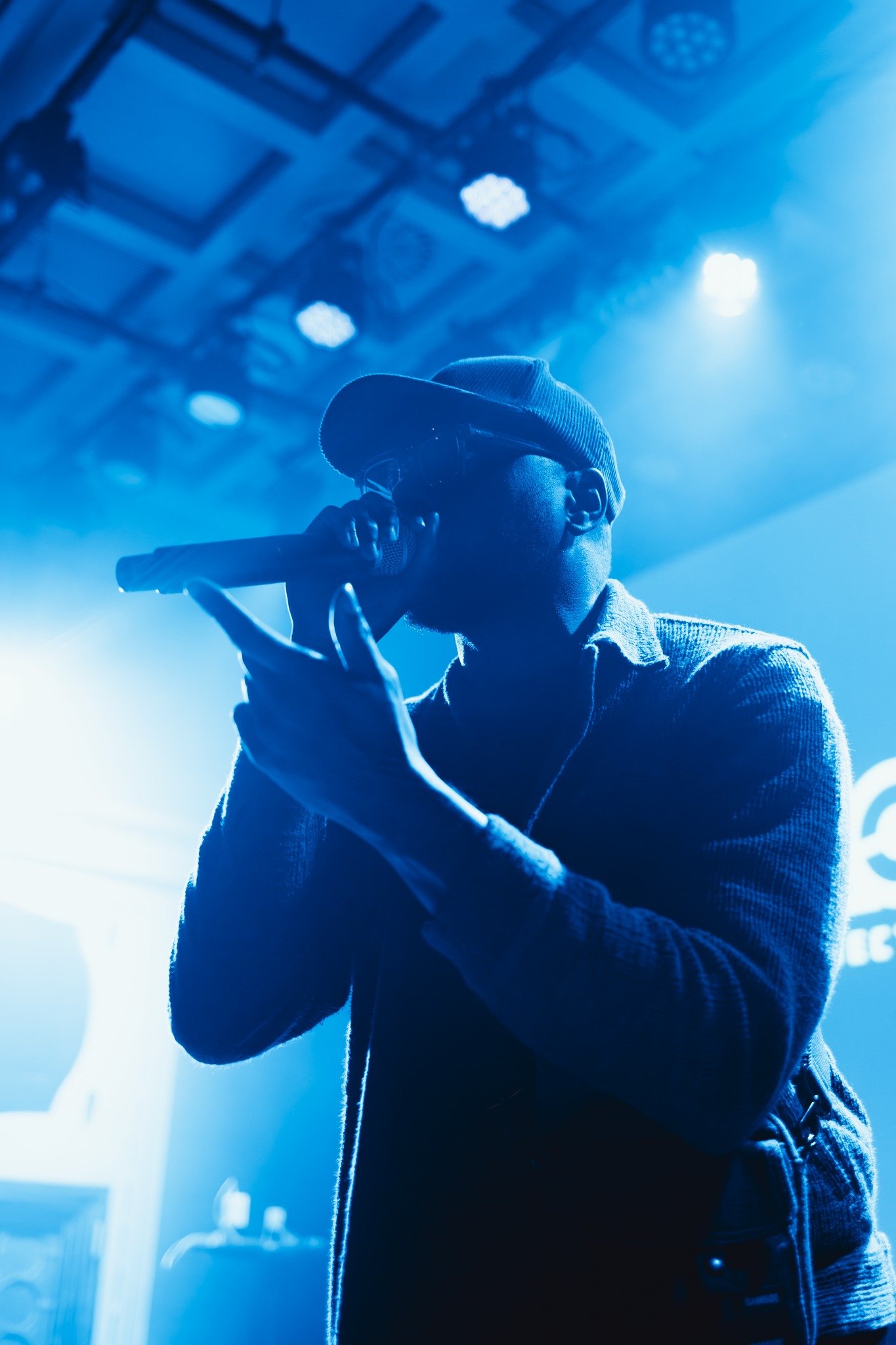 A silhouette of Kendan Adams performer singing into a microphone on stage, illuminated by blue stage lights, with visible stage equipment in the background.