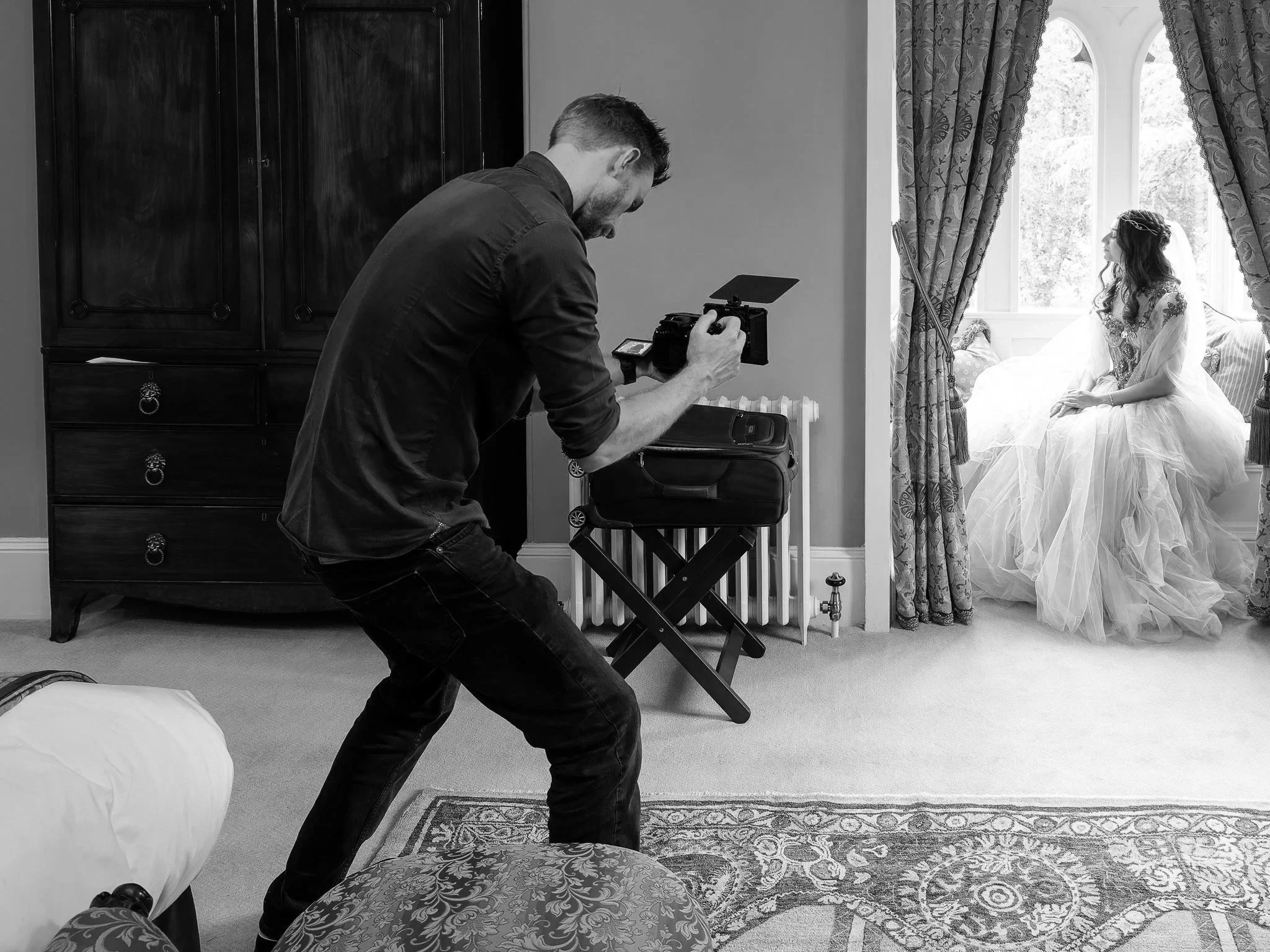 A videographer takes pictures of a woman in a wedding dress sitting by a window.