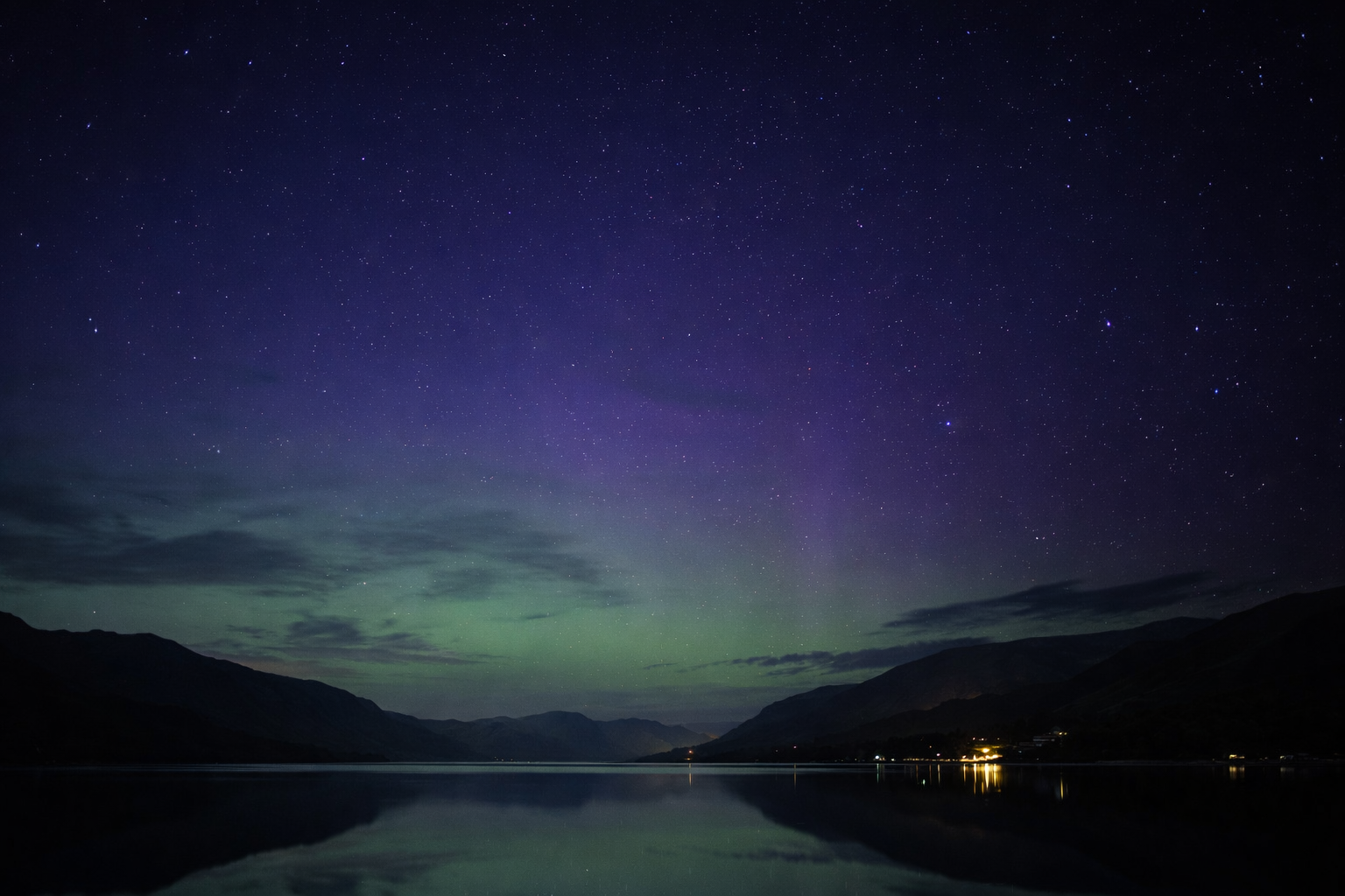 Nighttime landscape with a star-filled sky, the Aurora Borealis visible over mountains, reflected on a calm body of water.