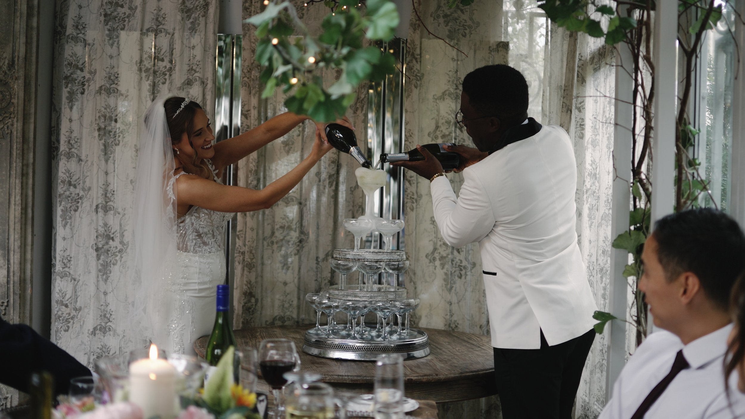 Bride and groom pouring champagne into a pyramid of glasses at their wedding celebration, with guests watching and a lit candle on the table.