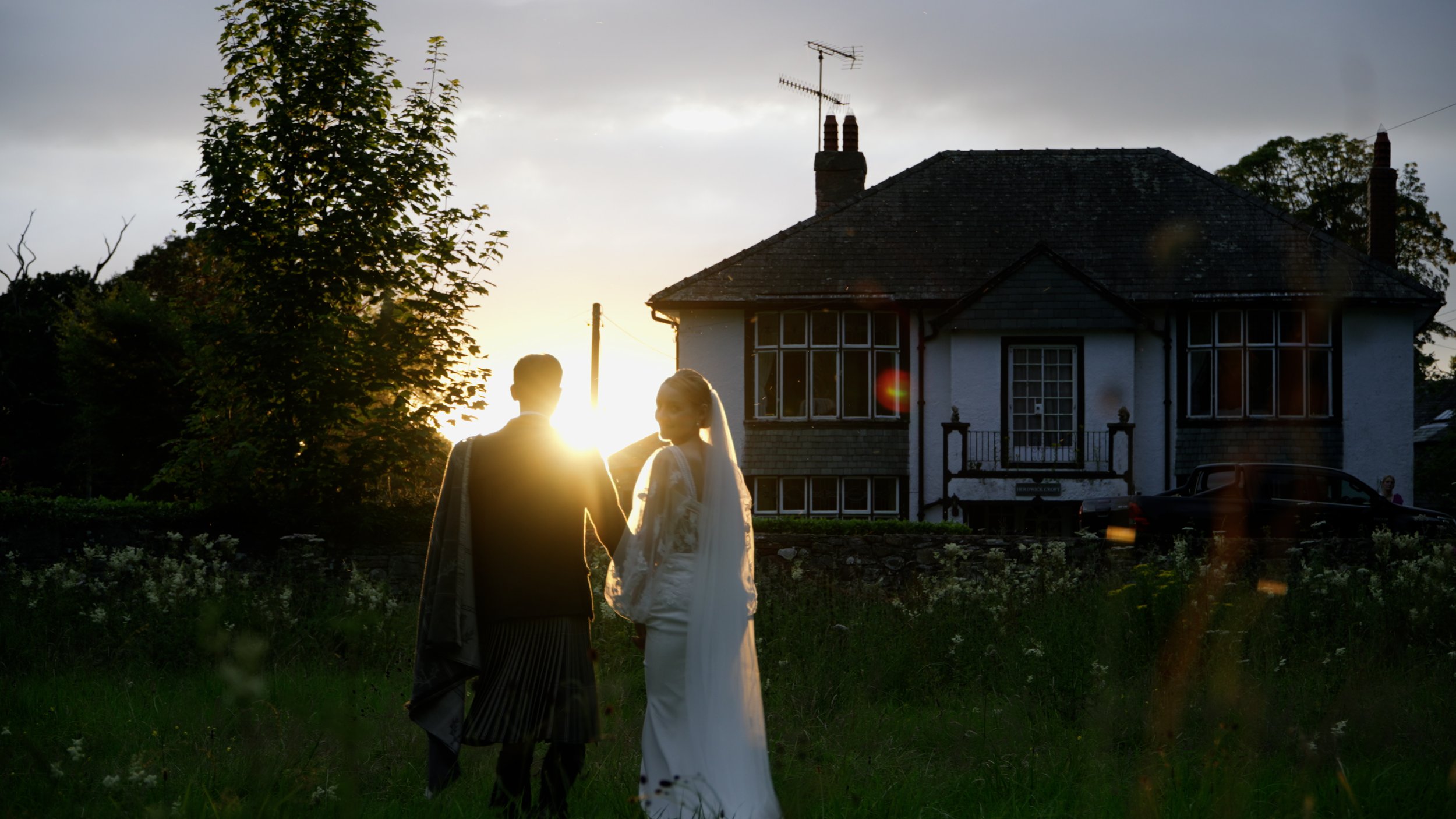 A bride and groom walking hand in hand outdoors at sunset, with a house and trees in the background.