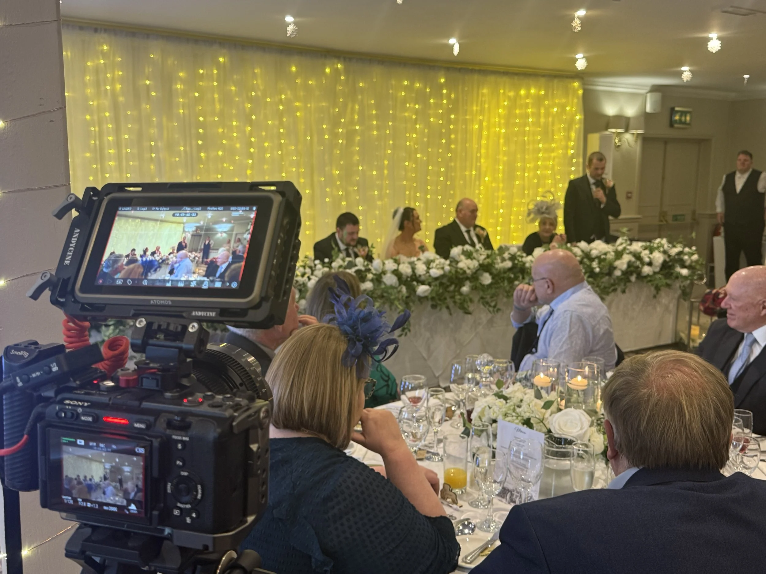 A wedding reception with a head table decorated with white flowers and backdrop of yellow fairy lights. Guests are seated at tables with floral centerpieces, and a man is giving a speech with a microphone.