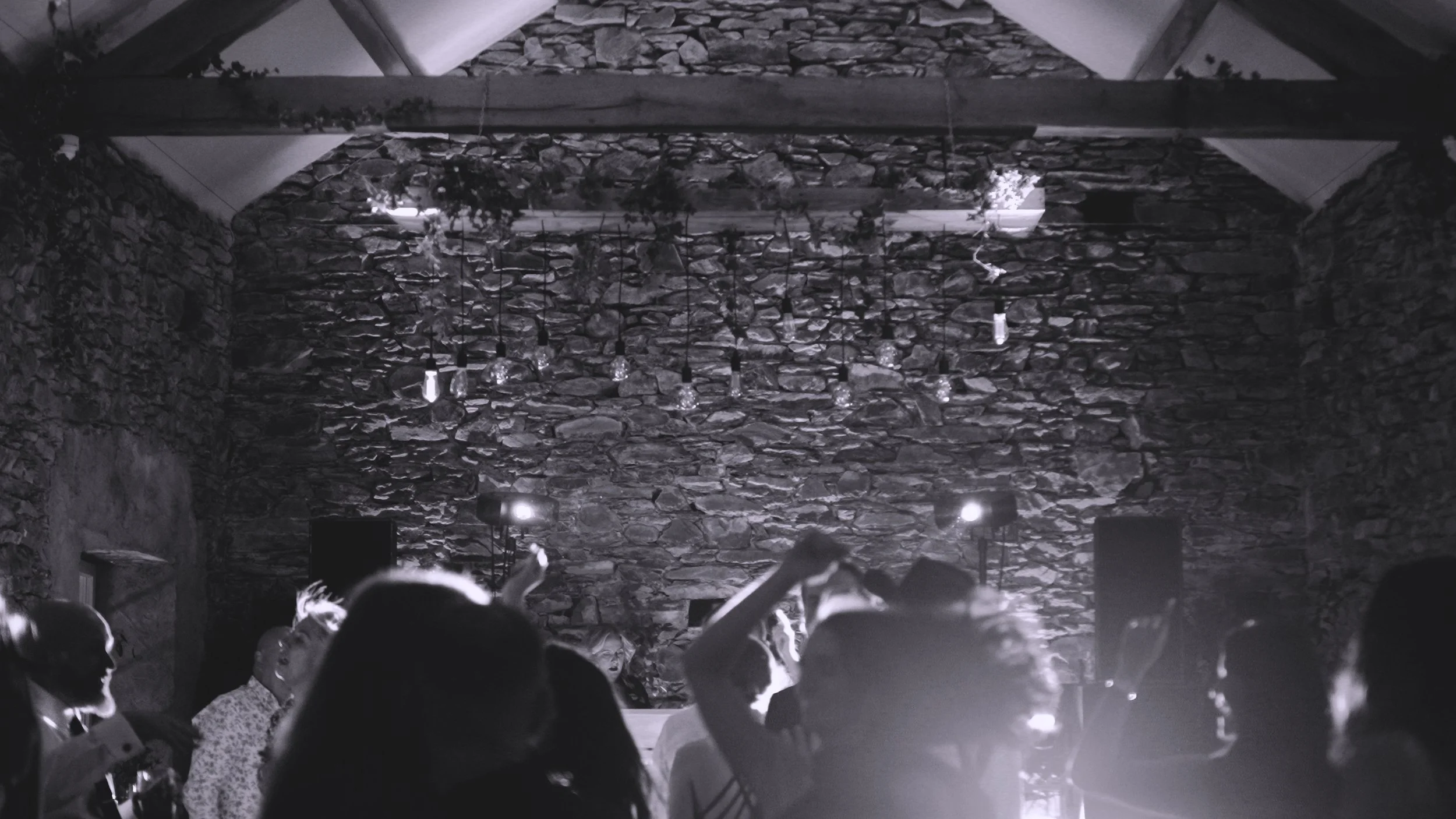 A black and white photo of people dancing in a rustic indoor venue with a stone wall backdrop and exposed ceiling beams, illuminated by string lights.