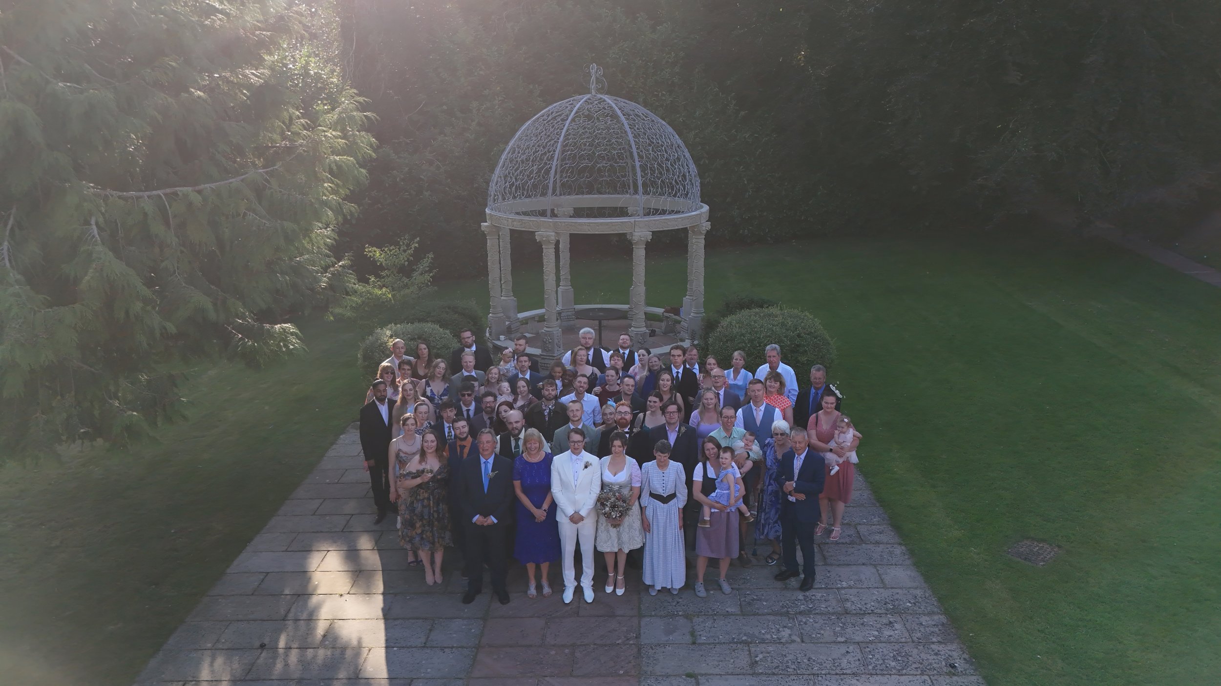 A large group of people in formal attire assembled outdoors for a photo, standing on a paved pathway in front of a decorative gazebo in a garden with trees and greenery.