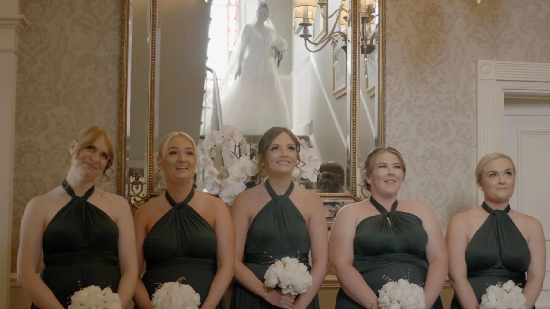 A group of five women in green dresses standing in front of a mirror, each holding a bouquet of white flowers. A bride in a wedding gown is reflected in the mirror, standing on the stairs behind them.