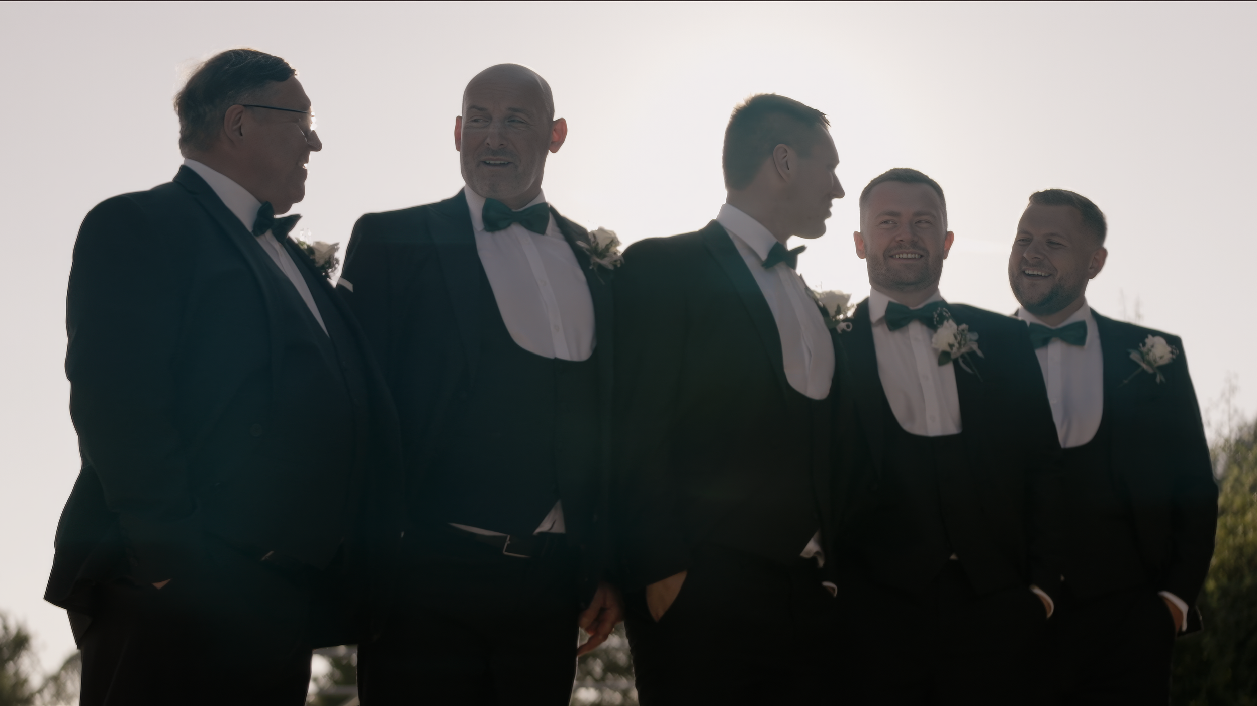 Five men in tuxedos with bow ties and boutonnières standing outdoors, talking, backlit by the sun.