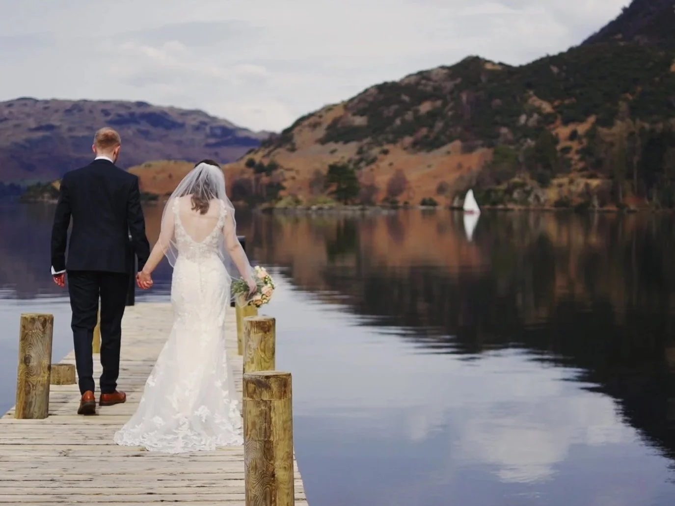 A bride and groom walking hand in hand on a wooden dock by a calm lake with hills in the background, with a sailboat on the water.