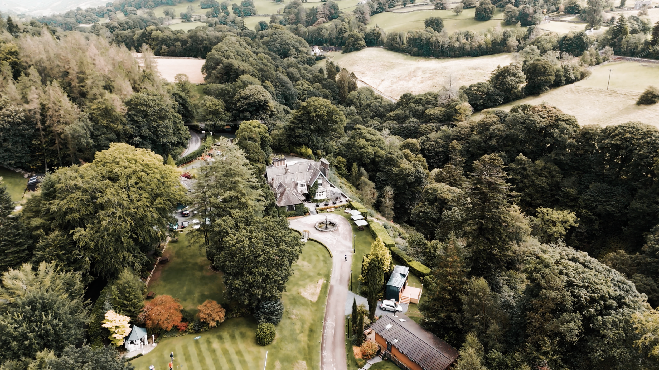 Aerial view of a large estate surrounded by lush forest with a house, driveway, fountain, and garden, set in a rural landscape with rolling hills and farmland in the background.