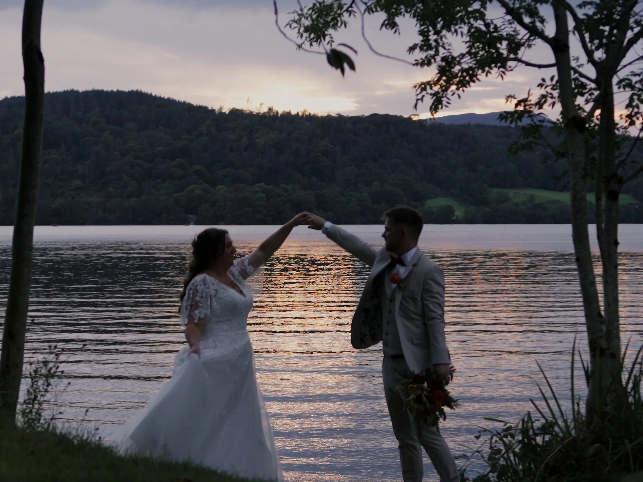 A bride in a white wedding dress and a groom in a light-colored suit dance by a lake at sunset, surrounded by trees and hills in the background.