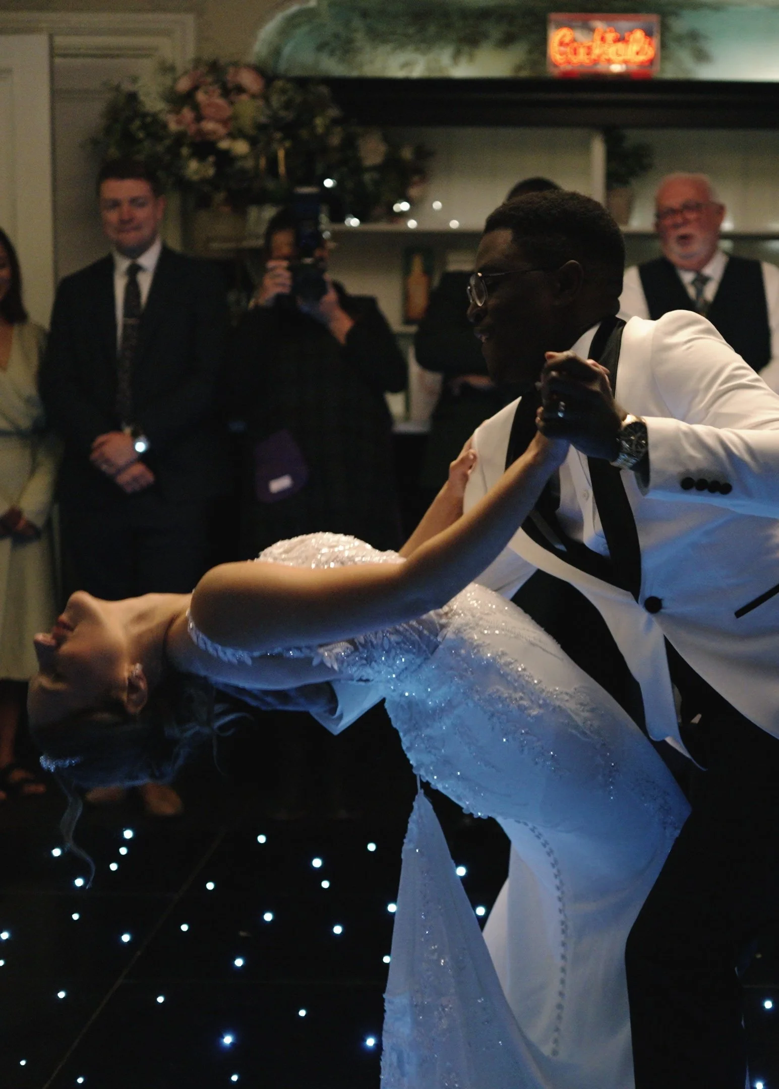 Couple dancing at a wedding reception, with the groom in a white tuxedo and the bride in a white wedding gown, surrounded by guests in formal attire watching and taking photos.