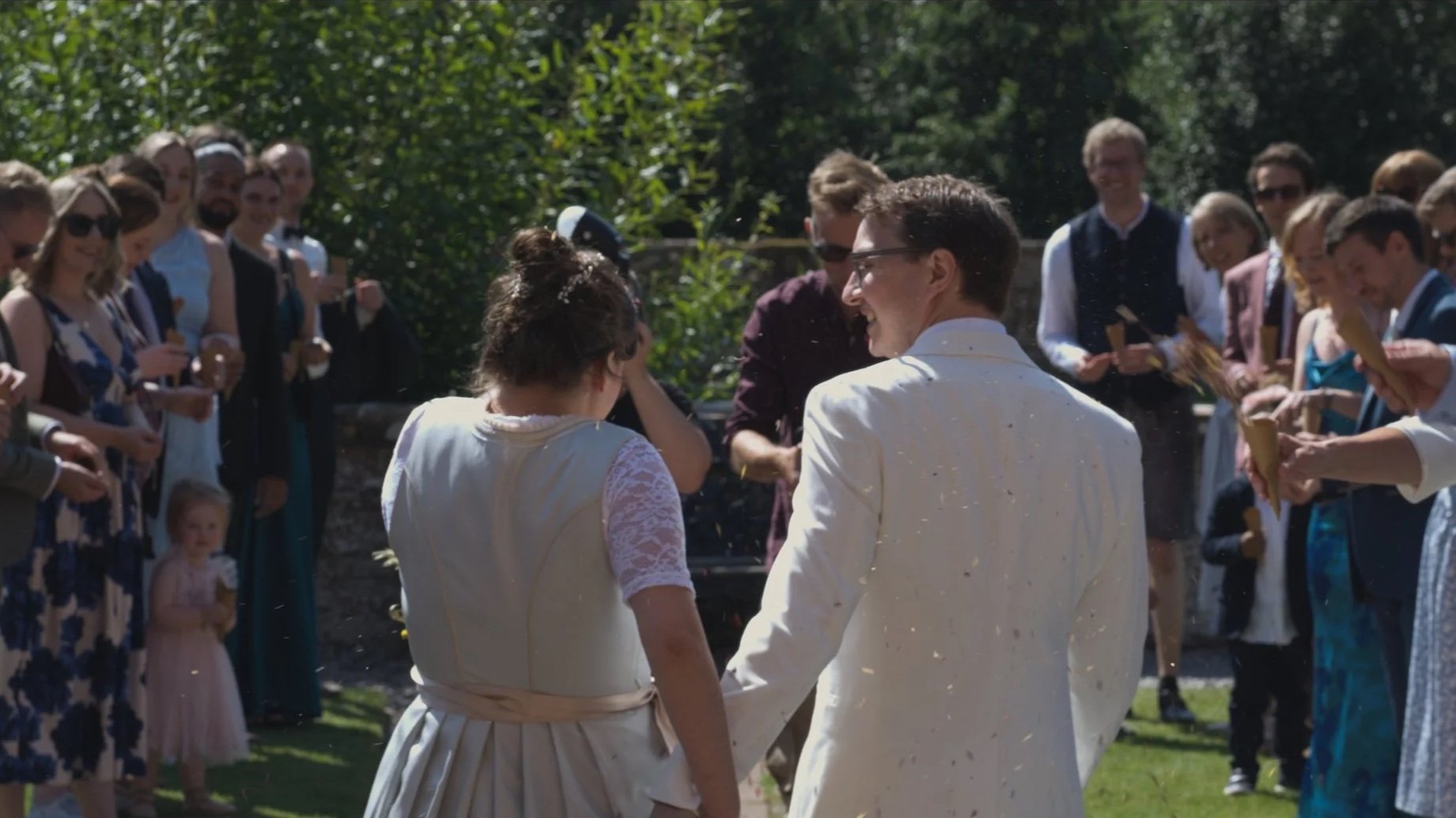 A wedding ceremony outdoors with a bride and groom holding hands, surrounded by guests. The bride is wearing a lace dress and the groom is in a white suit. The guests are dressed semi-formally and are watching the couple, some holding cones, on a sunny day with green foliage in the background.