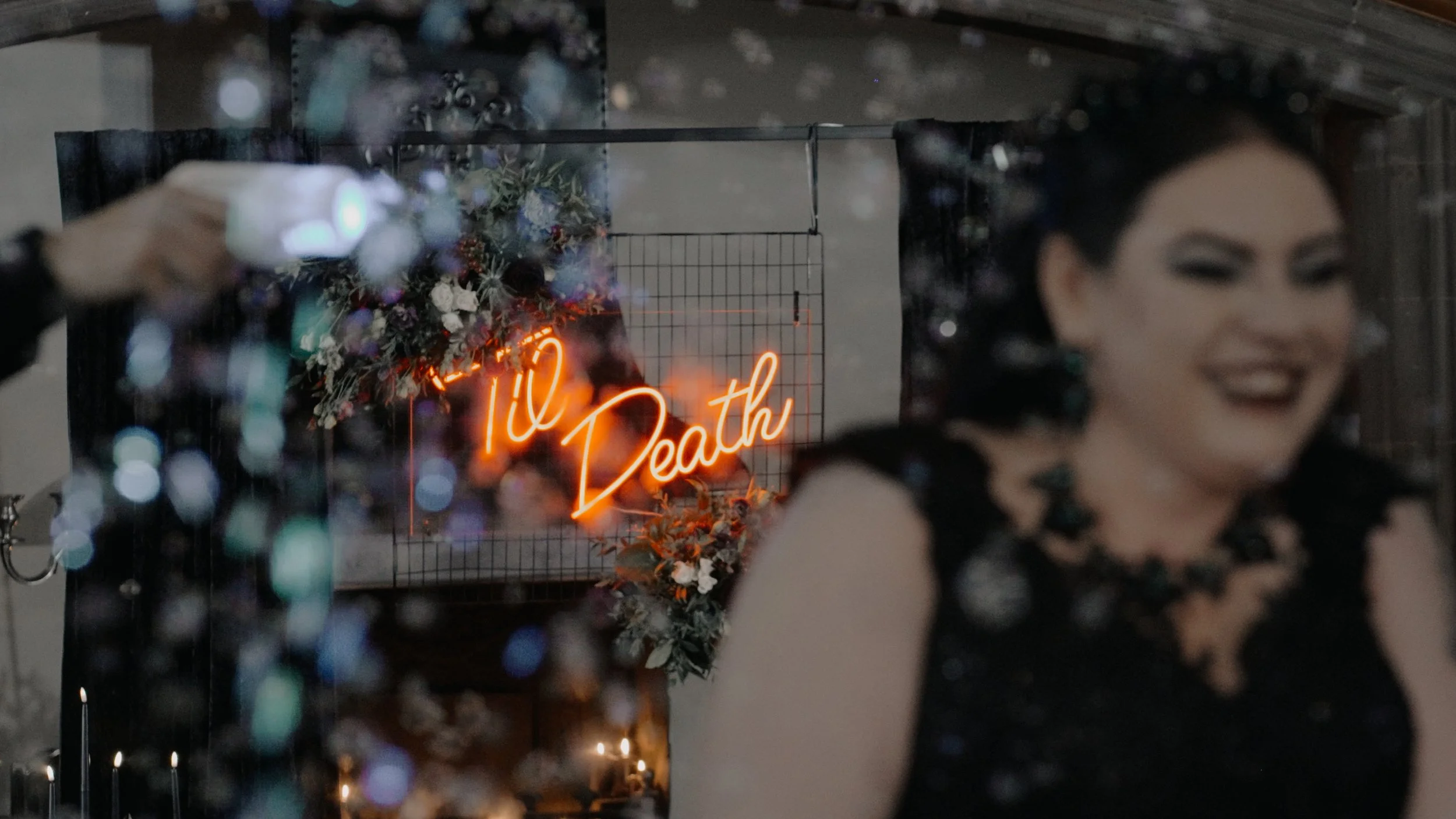 A woman with dark hair and makeup, dressed in black, is laughing as she walks down the aisle. In the background, there is a neon sign that reads 'til Death' and some decorative flowers.
