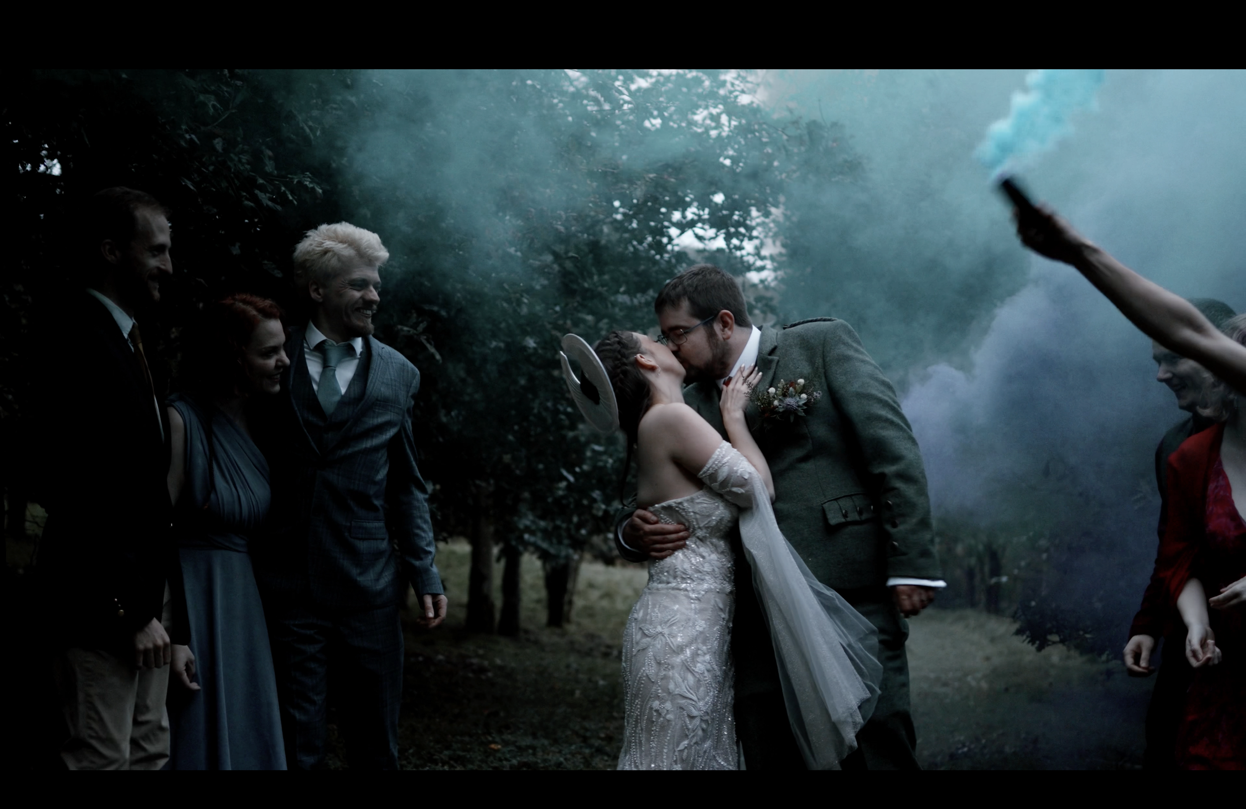A couple in wedding attire kissing outdoors, surrounded by friends holding smoke bombs, with trees in the background.
