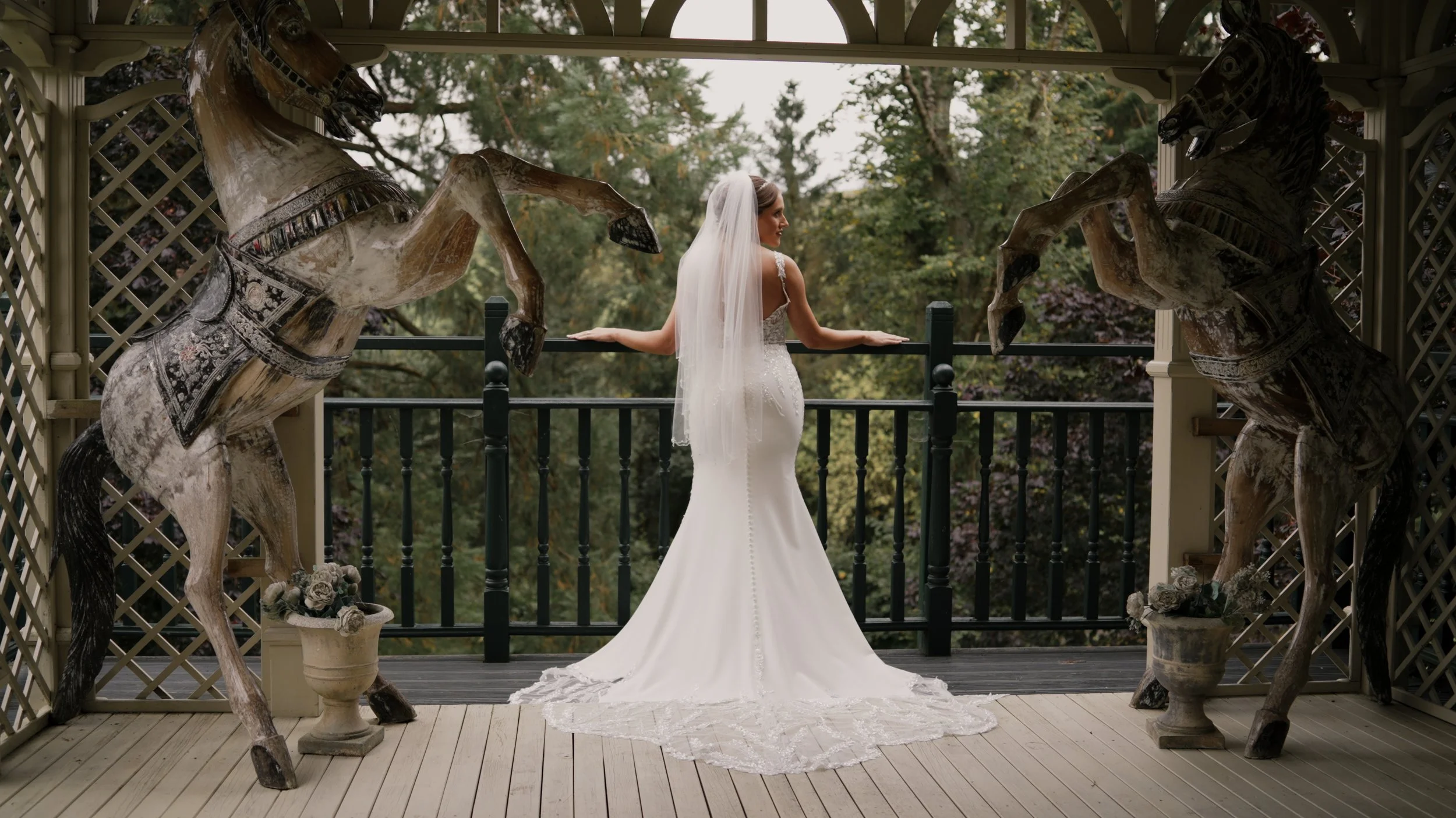 A bride in a white wedding dress with a long train and veil, standing on a porch with her back turned. Two large vintage carousel horses, painted white with black and silver accents, flank her on either side, each in a rearing position on stone pots 