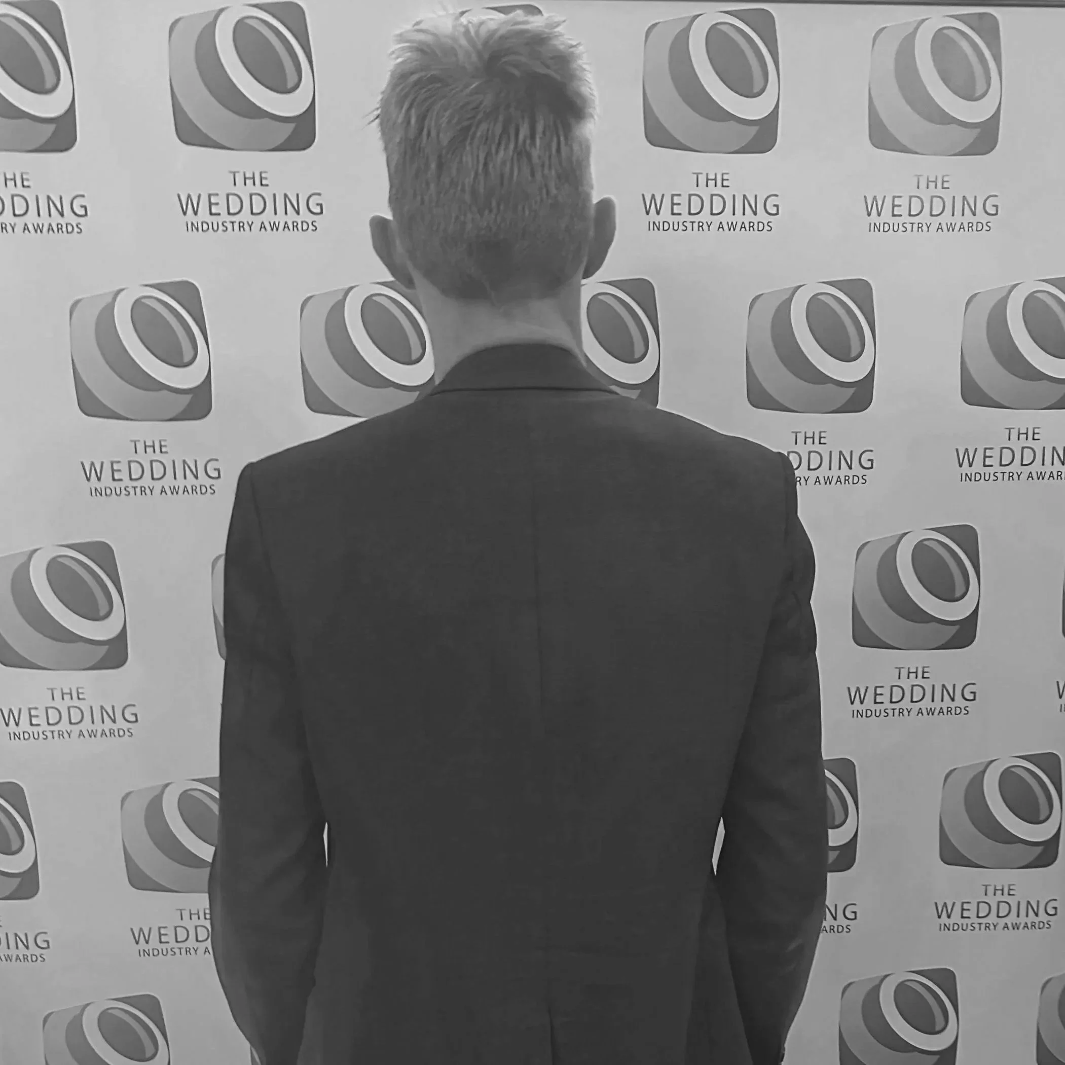 Graham Wilcock standing with his back facing the camera, in front of a backdrop with a logo and text for 'The Wedding Industry Awards'.