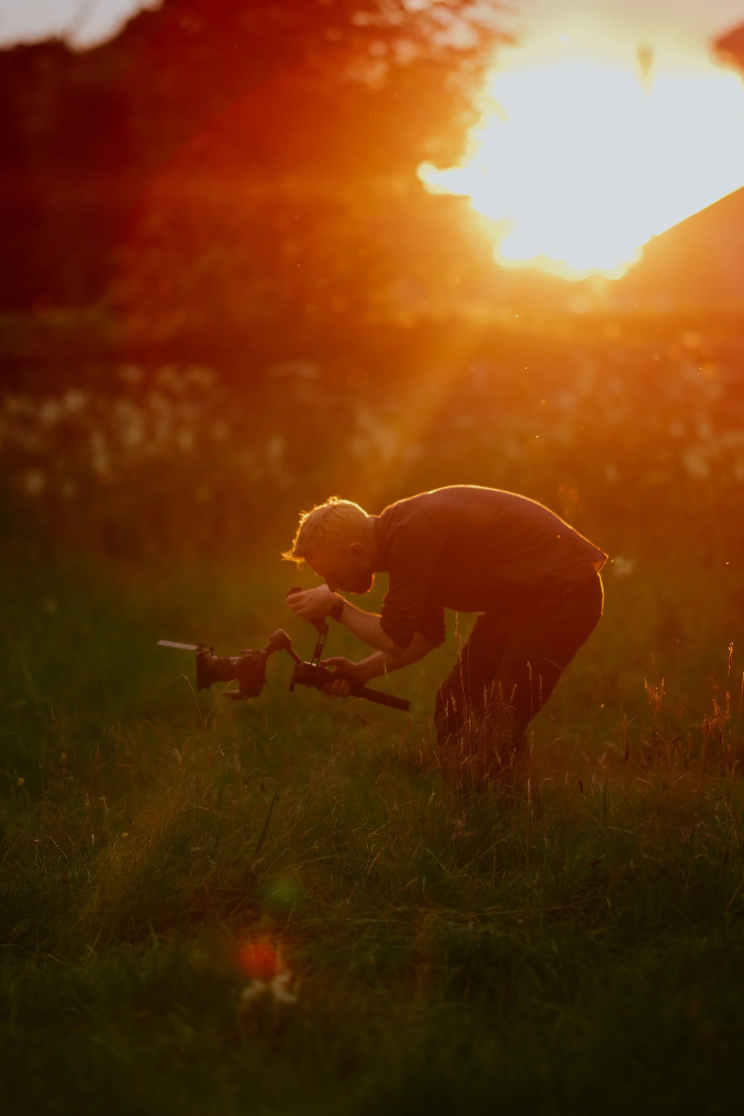 A person with light hair and a beard, wearing a dark shirt, bent over on a grassy field working with a camera stabilizer during sunset.