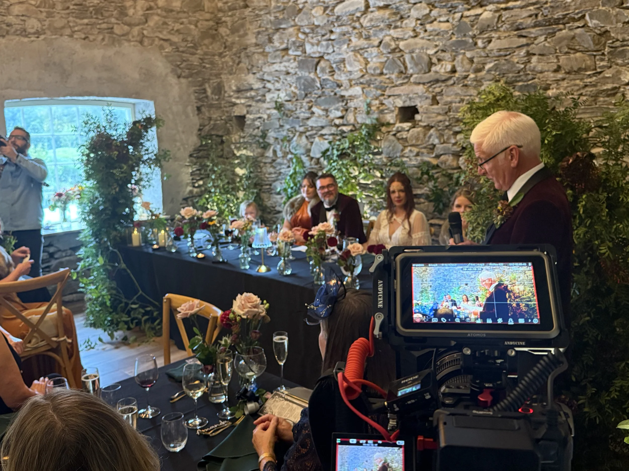 A wedding reception with a man giving a speech at a table decorated with flowers, with guests seated and a camera recording the event in a rustic stone-walled venue.