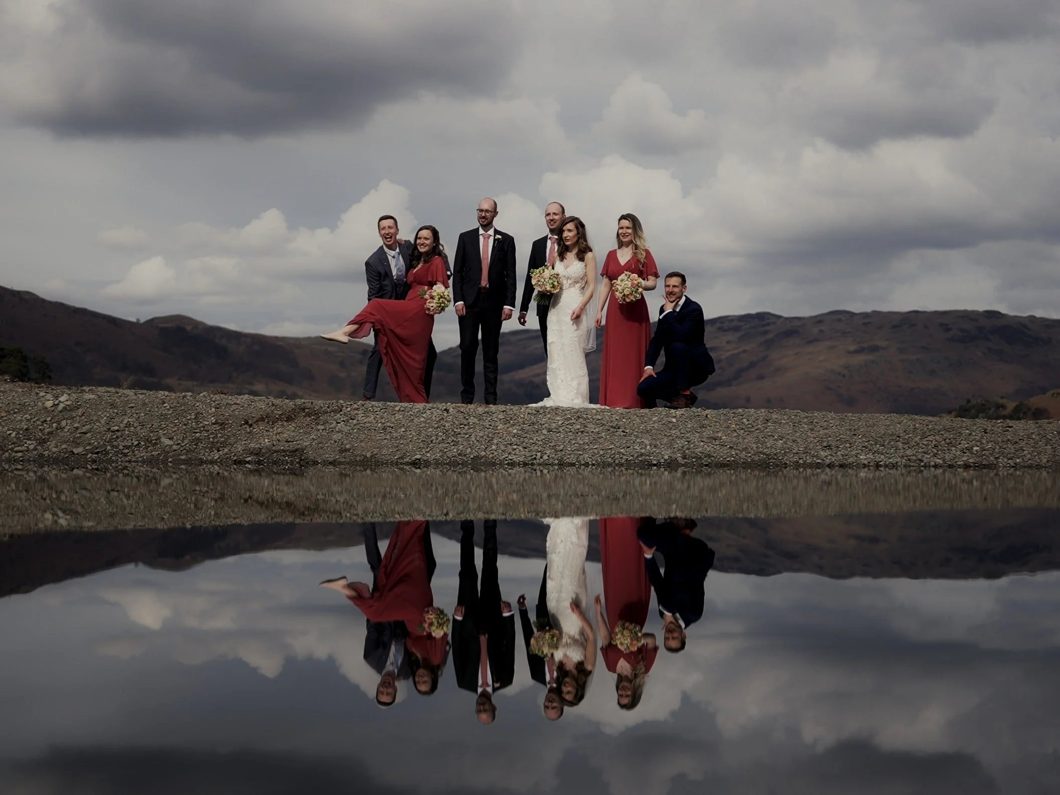 Group of seven people, including a bride in a white wedding dress and bridesmaids in red dresses, posing outdoors on rocky terrain with a mountain landscape and cloudy sky in the background, reflected in a body of water.