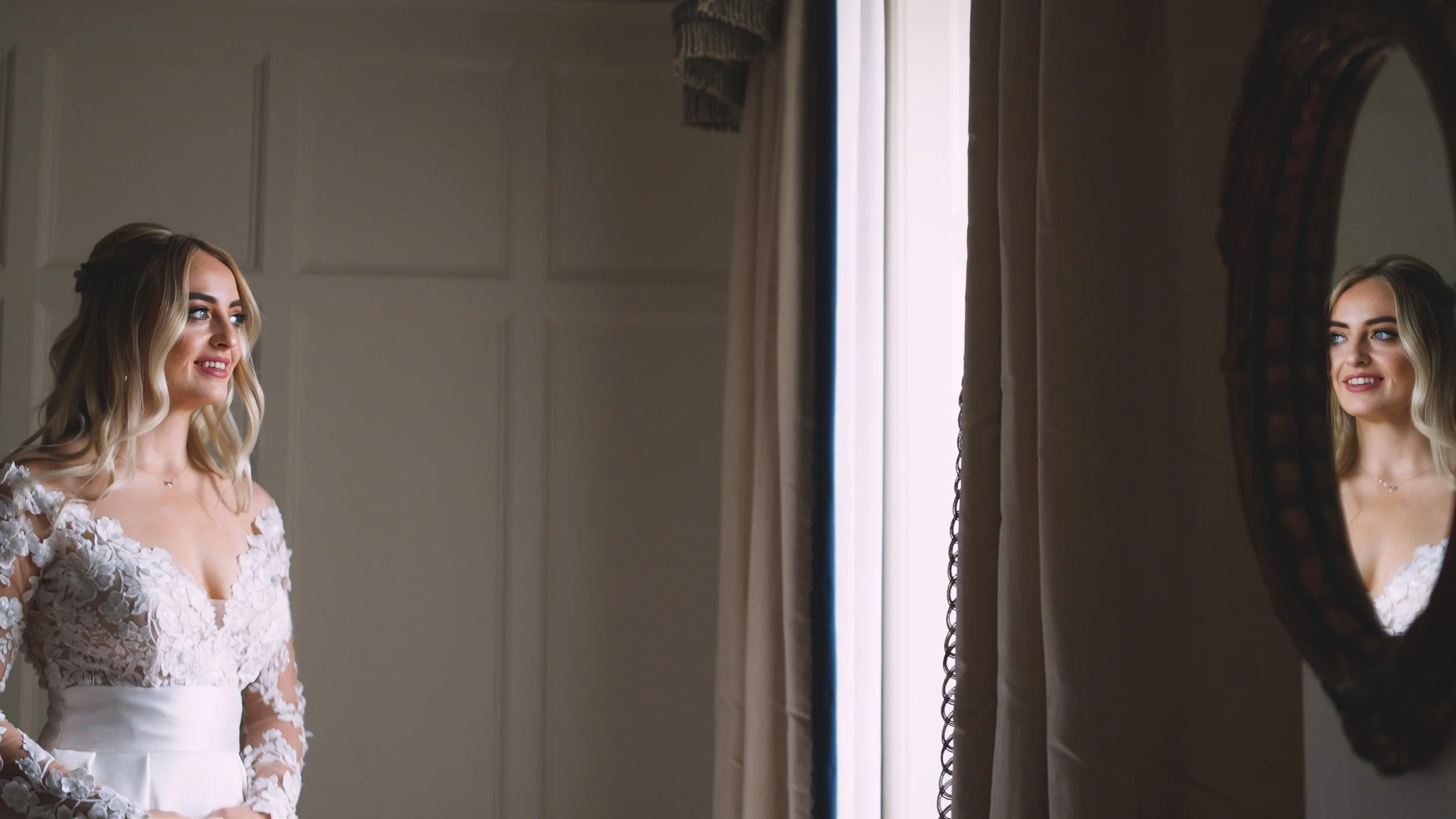 A bride in a white wedding dress with lace details stands and smiles while looking at her reflection in an oval mirror.