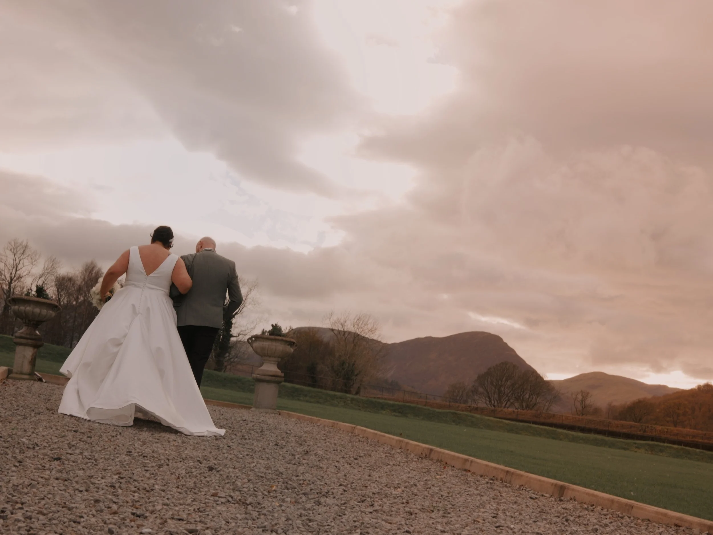 A bride in a white wedding dress walking arm-in-arm with a groom in a grey suit outdoors during sunset with a mountainous landscape and cloudy sky in the background.