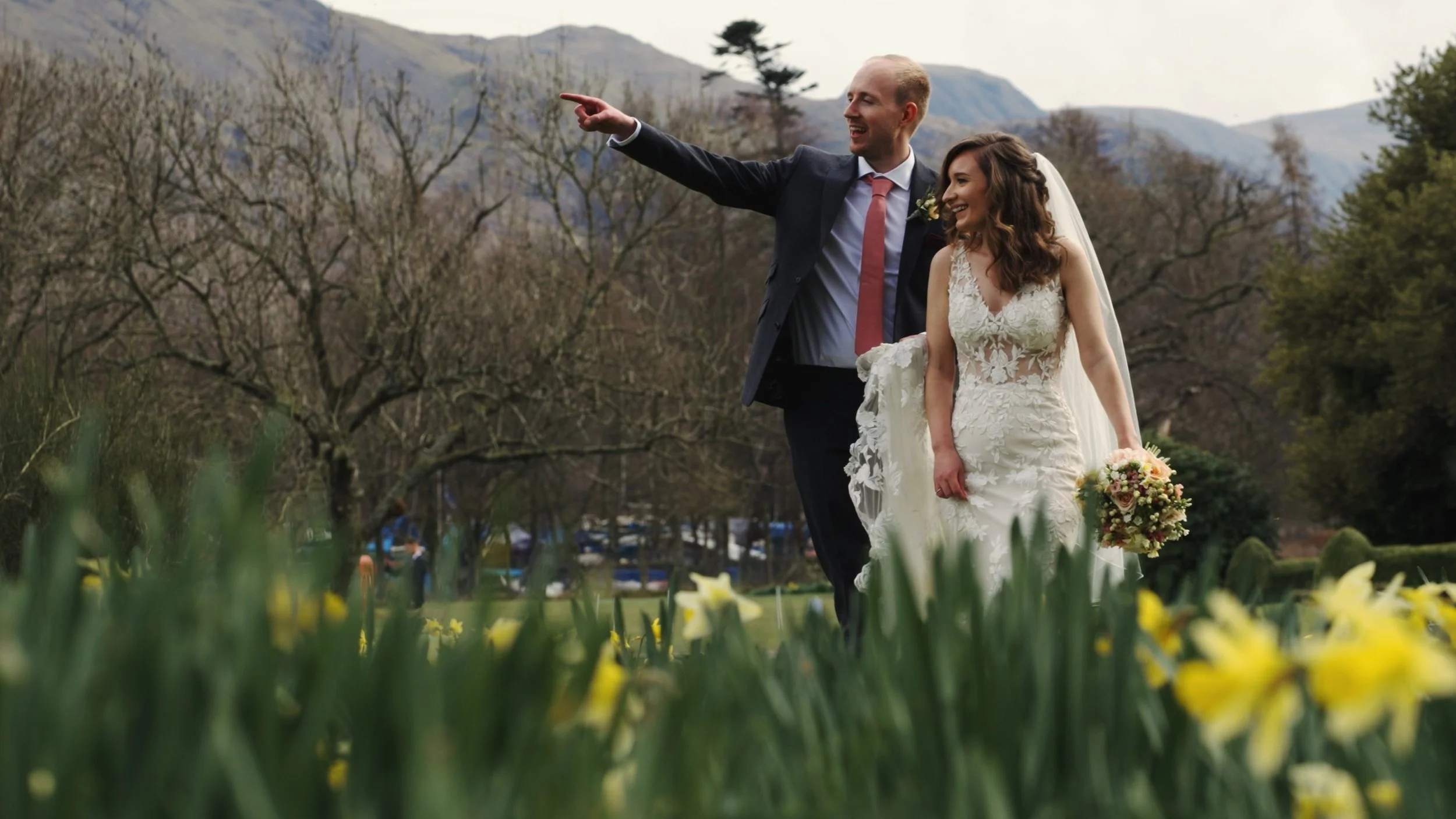 A bride and groom walking outdoors in a park-like setting during their wedding, with the groom pointing at something in the distance and both smiling. The bride is holding a bouquet of flowers, and the background includes trees, mountains, and a clou