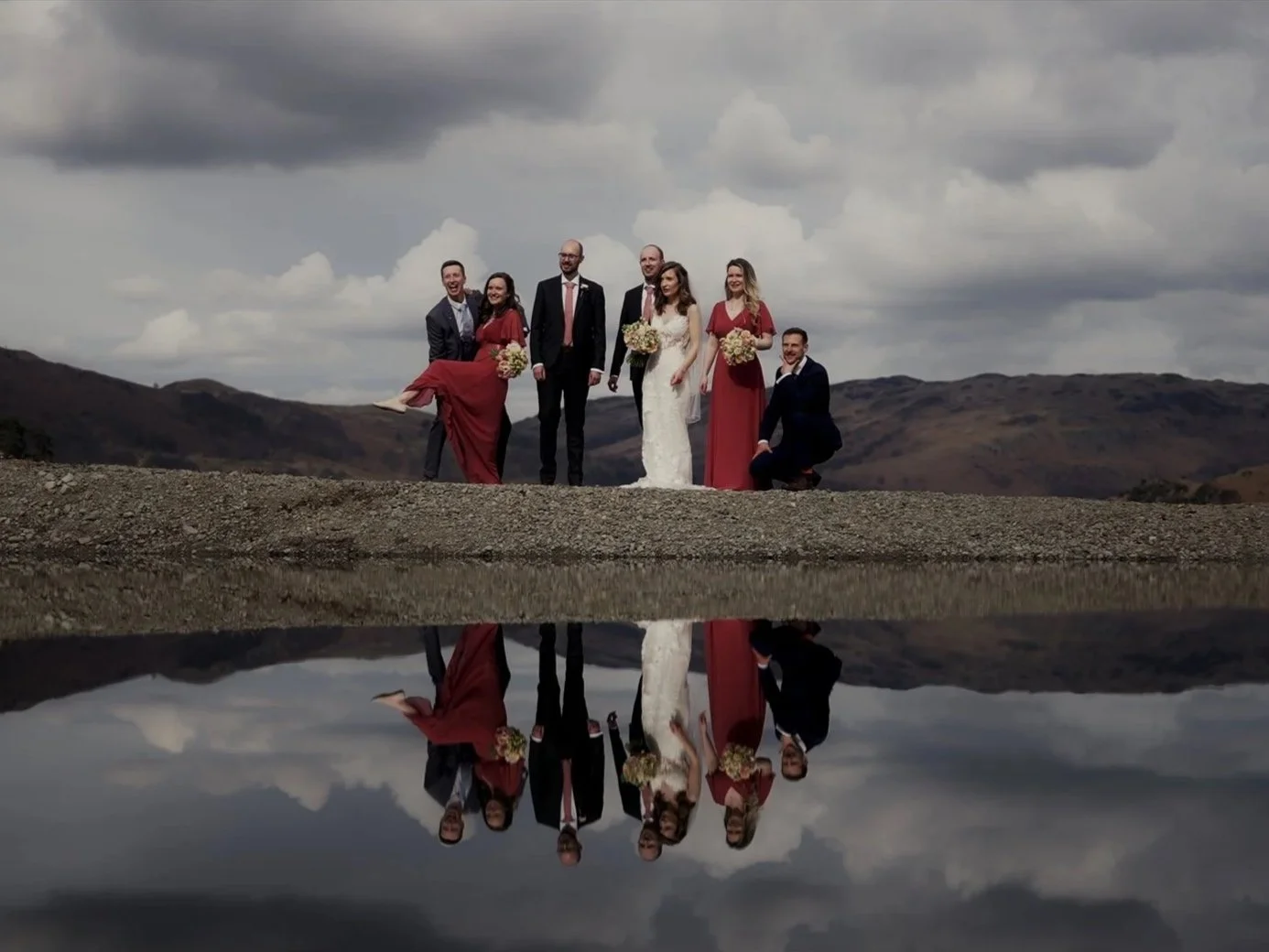 A group of seven people, including a bride in a white gown and five bridesmaids in red dresses, with two groomsmen, standing outdoors on rocky terrain under cloudy skies, with their reflection visible in a body of water.