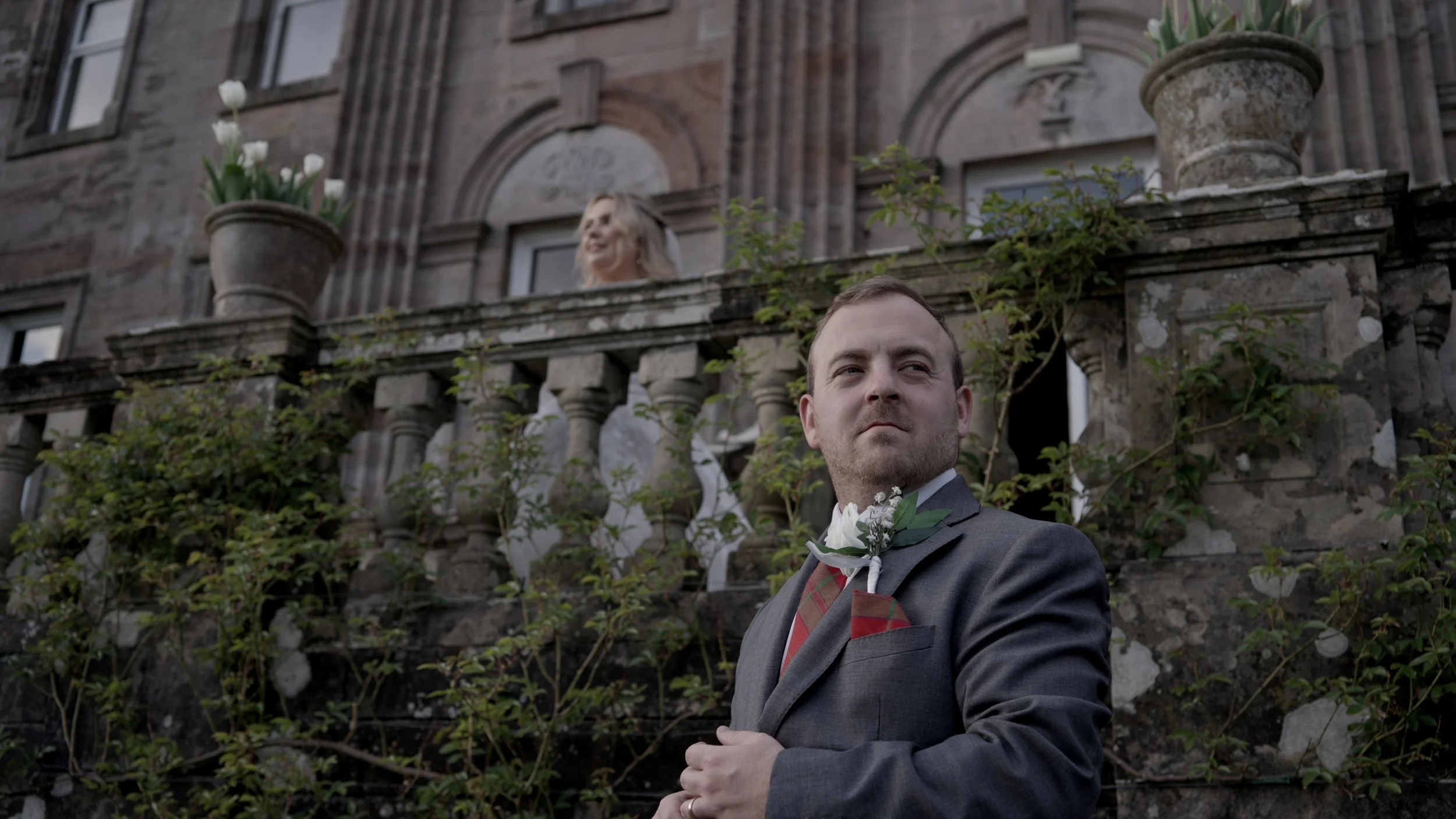 A man dressed in a suit with a boutonniere stands outside in front of an old stone building with a balcony, looking thoughtfully to the side. A woman with blonde hair is on the balcony behind him, looking out over the scene.