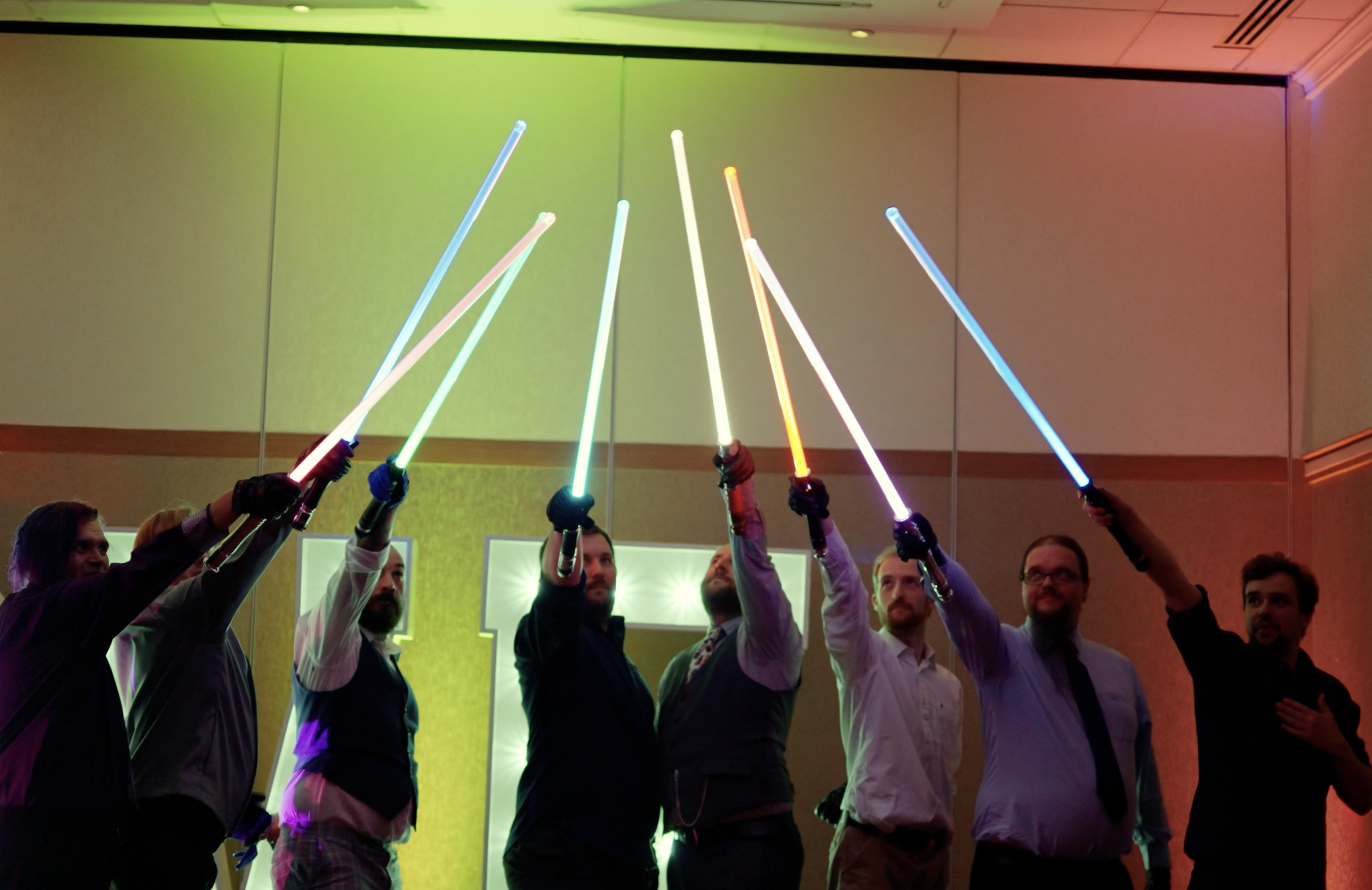 Group of six men dressed in formal attire holding illuminated lightsabers in a celebration or event setting.