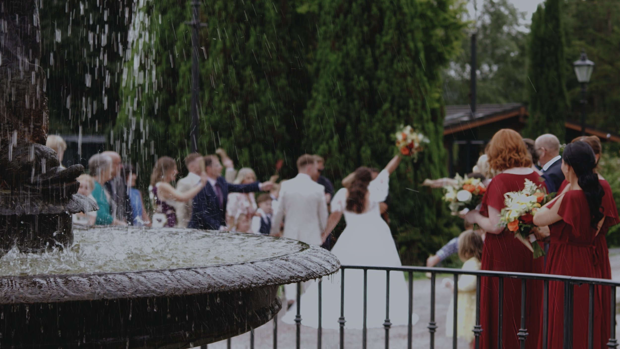 A wedding ceremony outdoors near a fountain, with guests and bridesmaids holding bouquets, rain falling, green trees, and a building in the background.