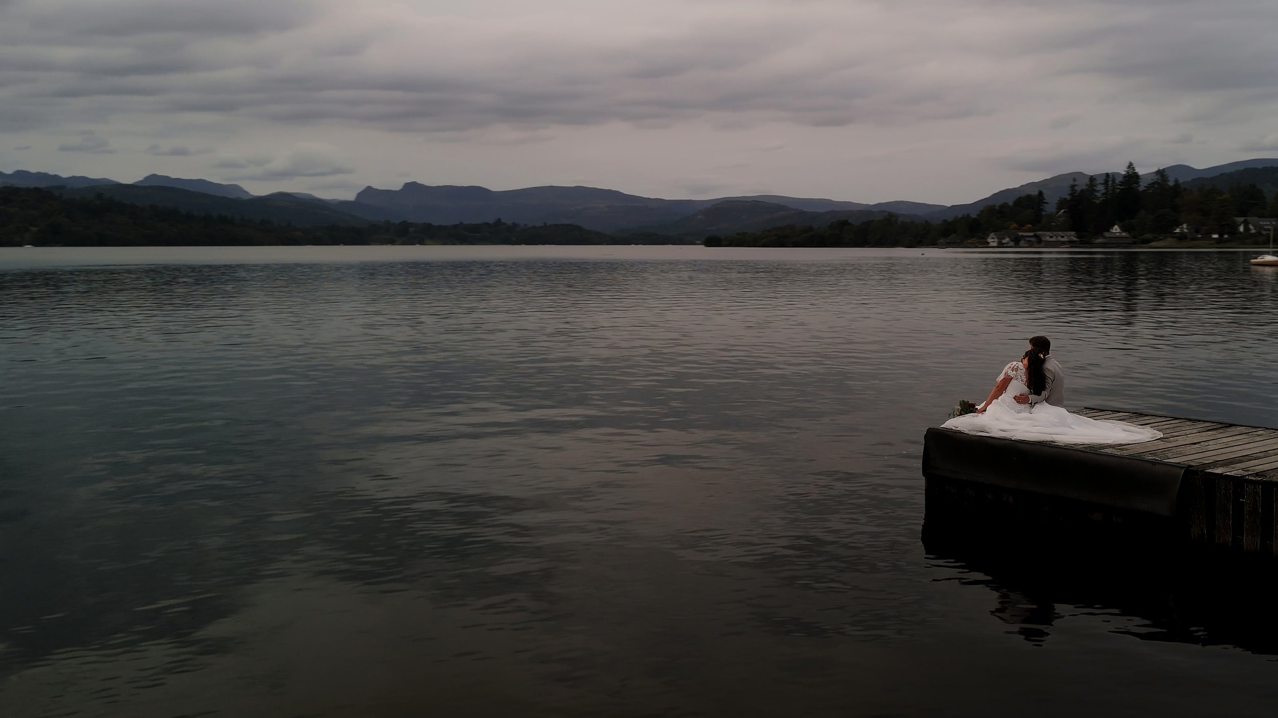 A bride in a white wedding dress sitting on a wooden dock by a calm lake, with mountains and cloudy sky in the background.