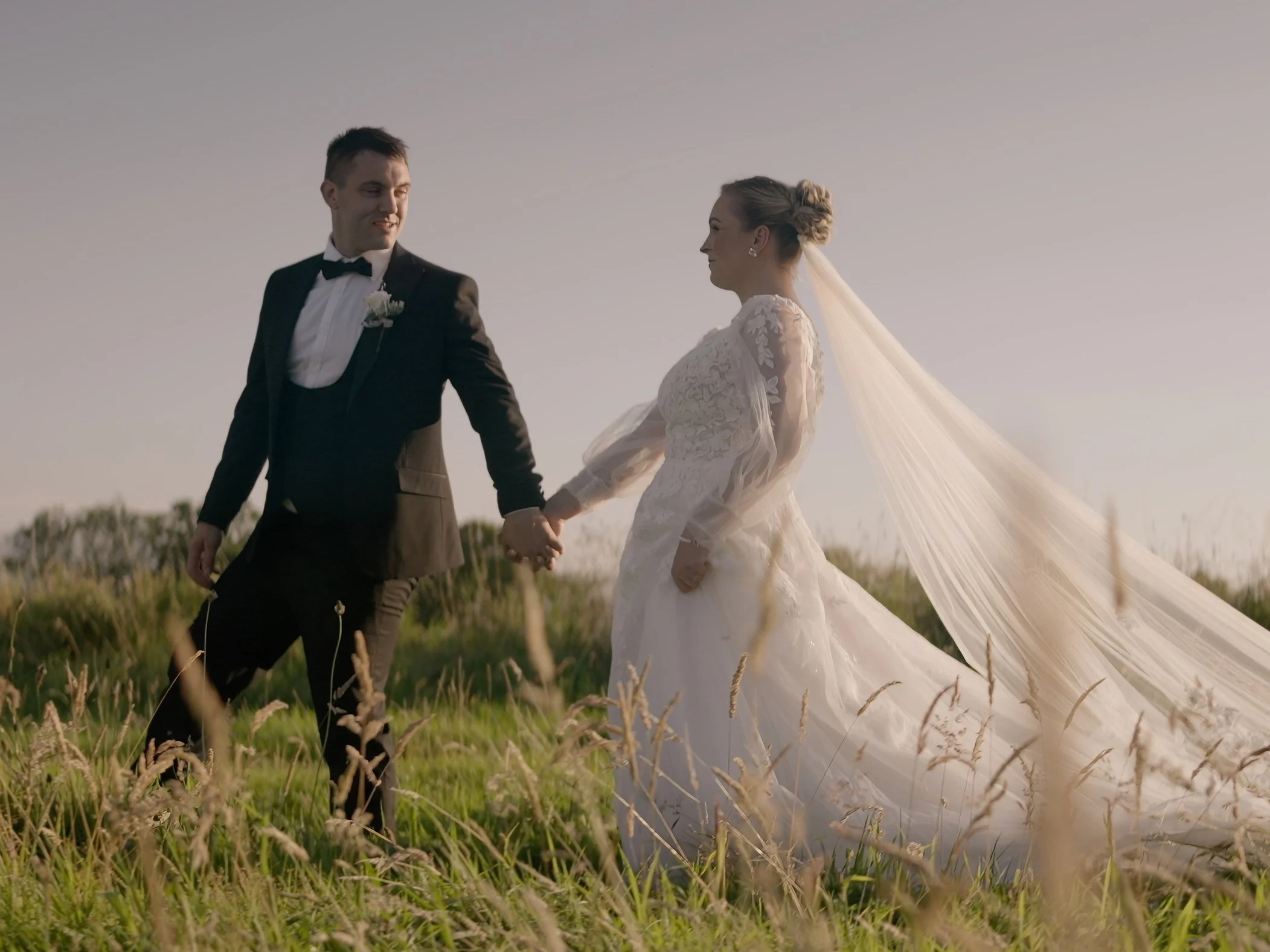 A bride and groom holding hands in a grassy field during sunset, with the bride in a long white wedding gown and veil, and the groom in a black tuxedo with bowtie.