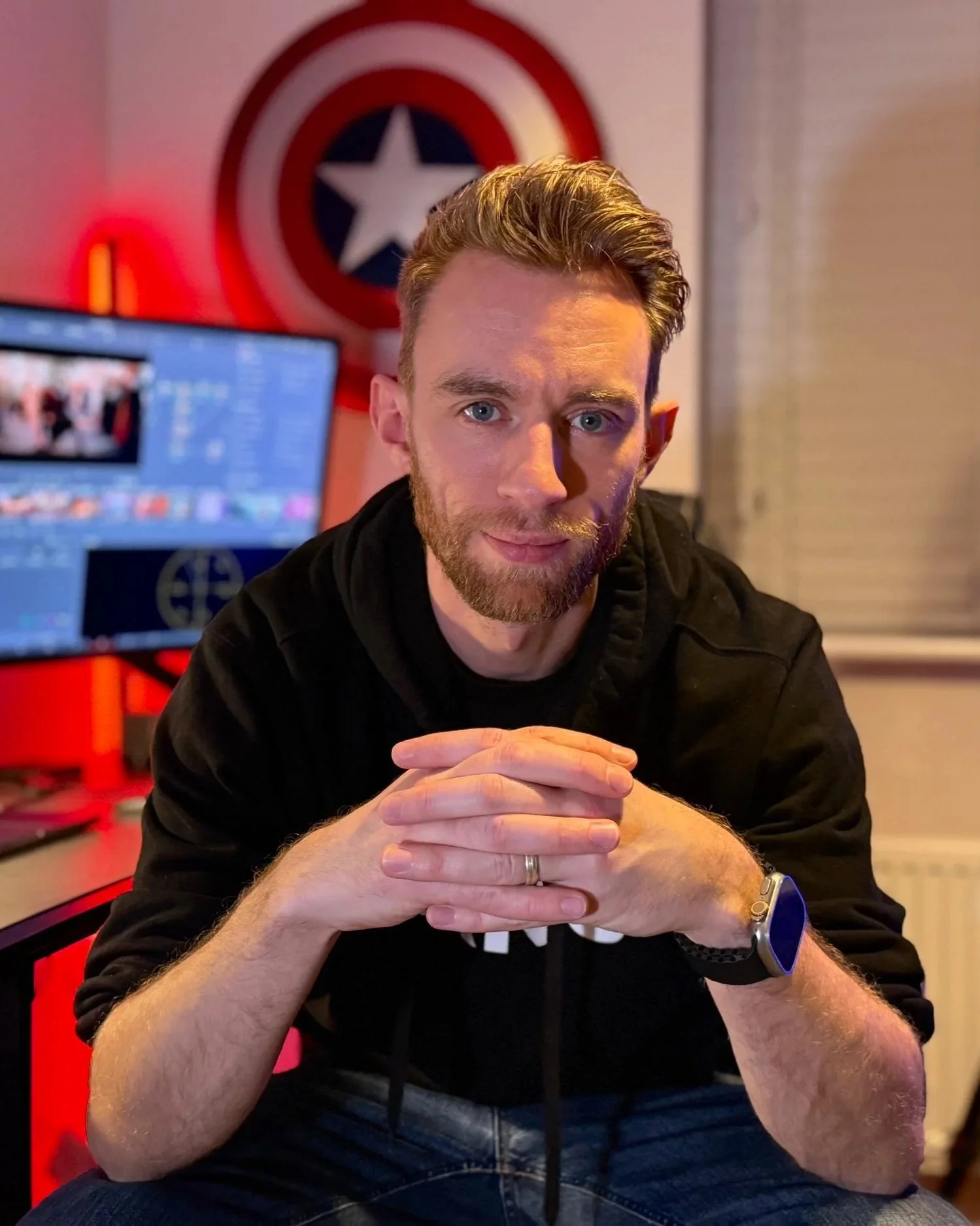A man with a beard and fair skin sitting indoors, with a shield resembling Captain America's behind him, a computer monitor with a video editing screen, and a window with closed blinds in the background.