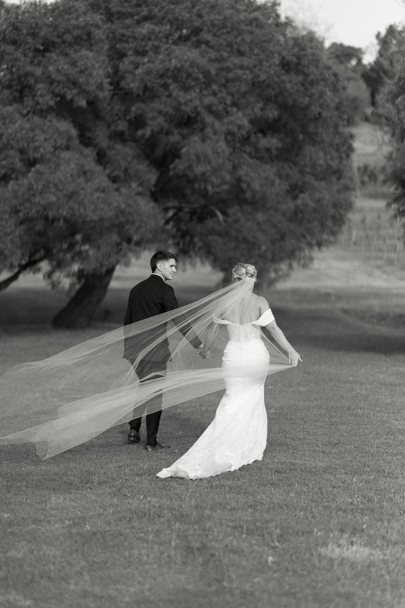 A black and white photo of a bride and groom holding hands and walking through a grassy park with large trees in the background.