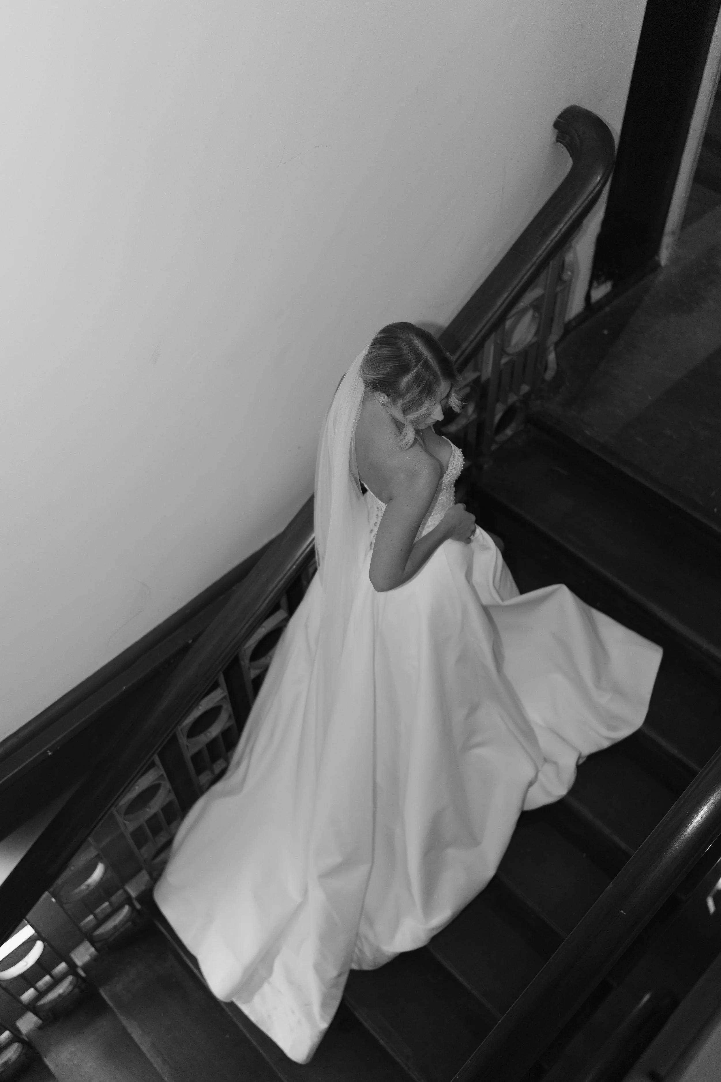 A bride in a wedding dress standing on a staircase, looking down, captured from above in black and white.