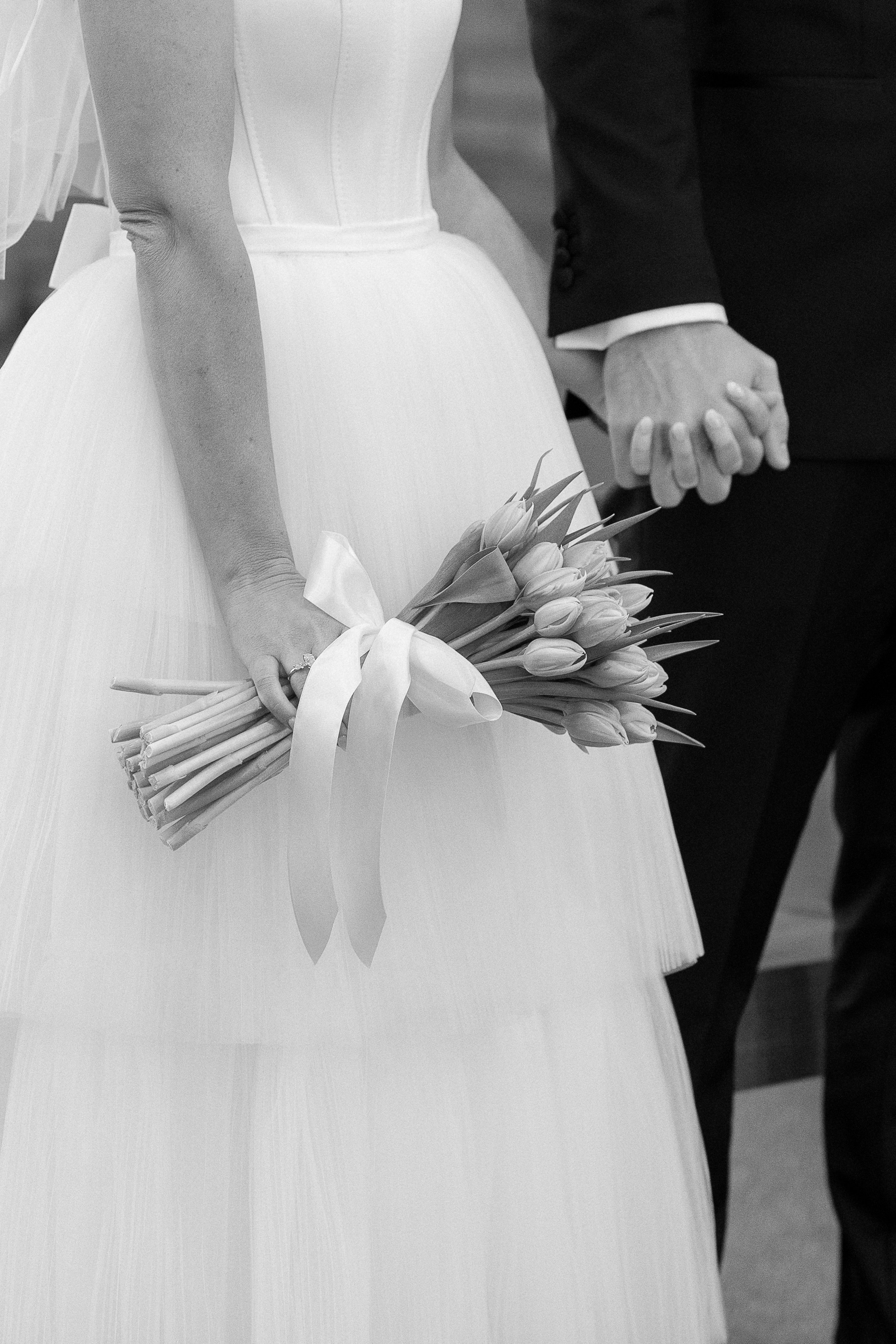 Bride holding a bouquet of tulips with a ribbon, standing next to the groom holding his hand during a wedding ceremony.