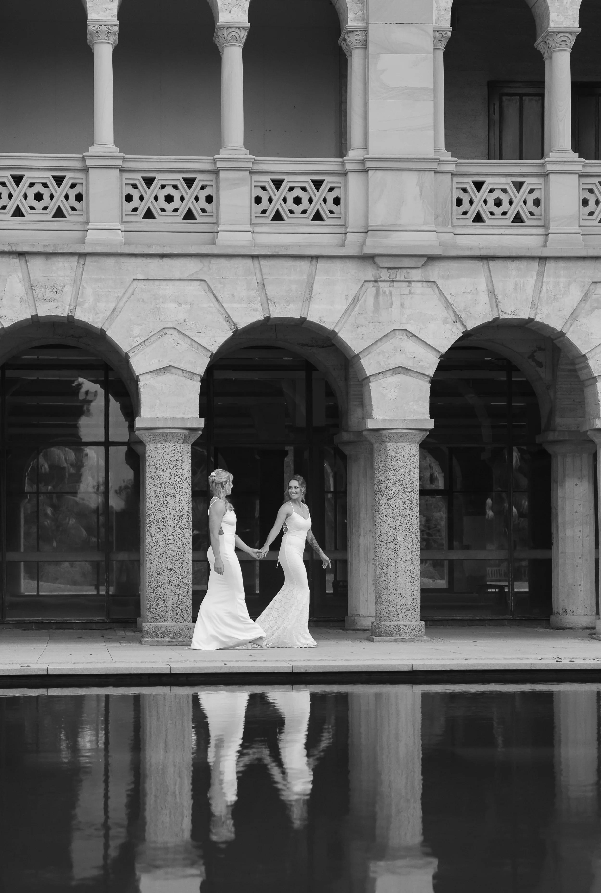 Two women in wedding dresses holding hands and walking beside a swimming pool with a stone building featuring arches and columns in the background.