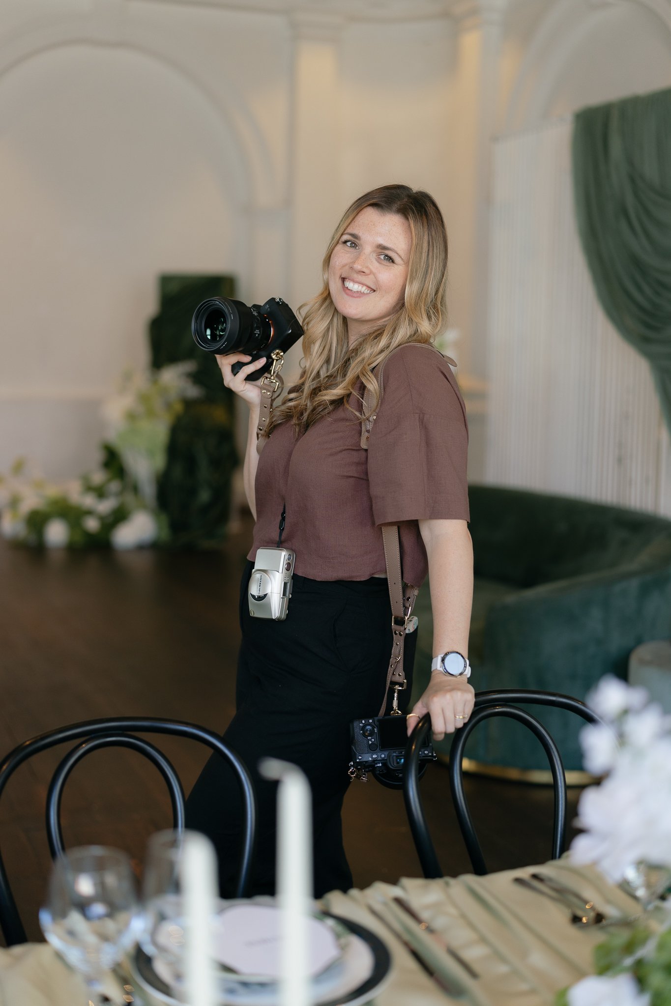 A woman with long wavy blonde hair smiling and holding a professional camera, standing in a decorated event space with tables, chairs, floral arrangements, and candles.