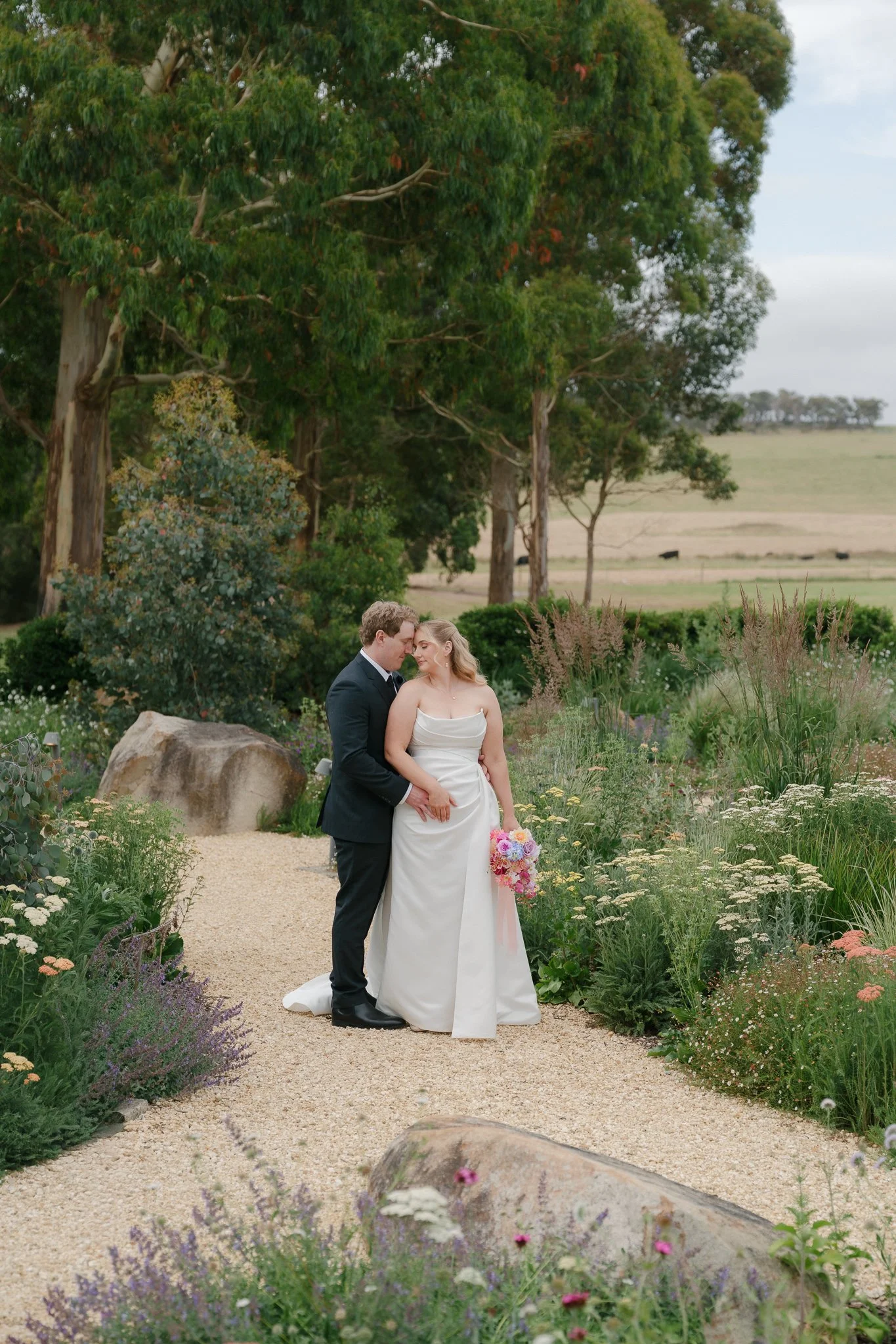 A bride and groom embrace on a gravel path surrounded by lush greenery and wildflowers during their wedding photo shoot.