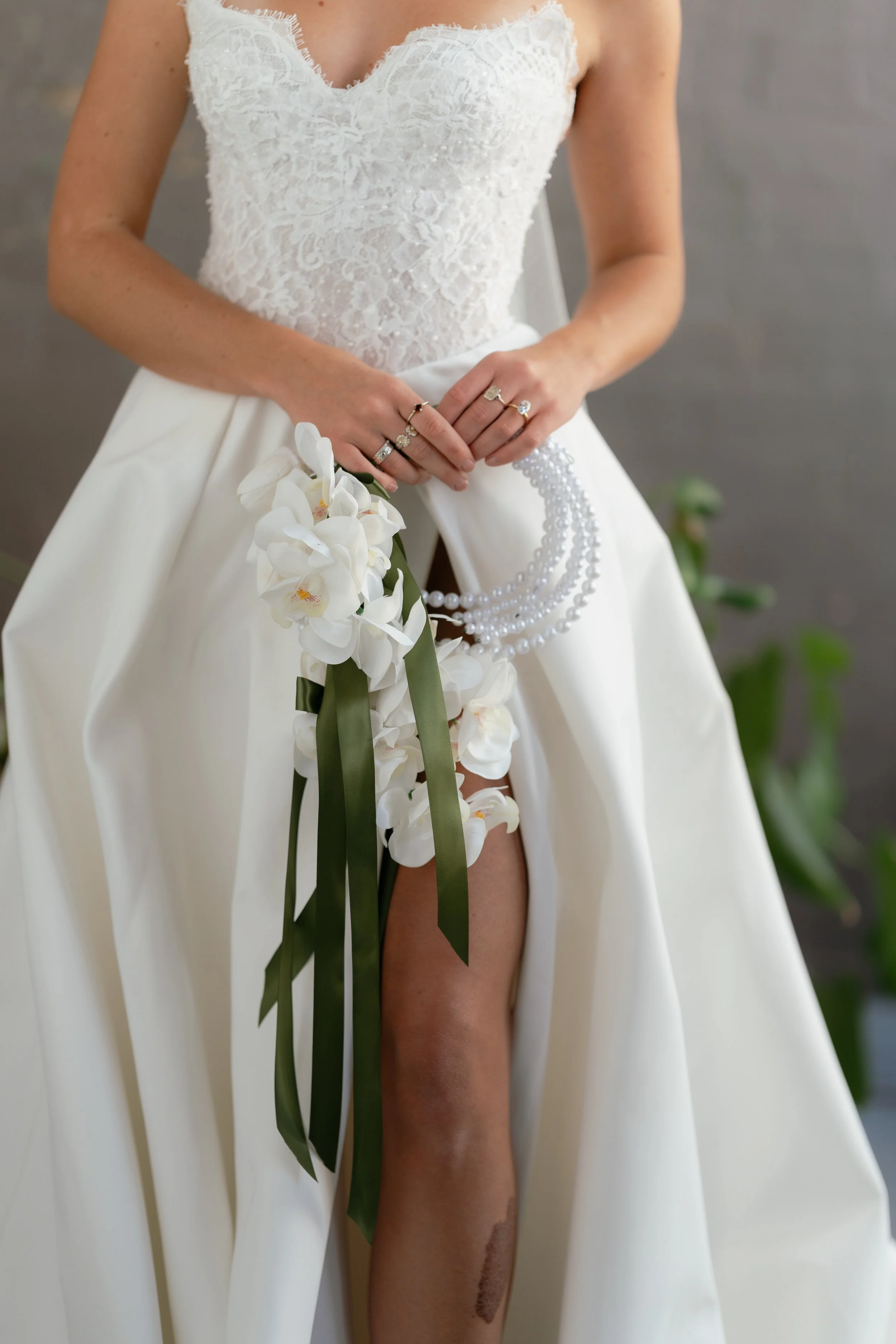 A bride wearing a lace wedding dress holding a bouquet of white orchids with long green ribbons, standing in front of a neutral background.