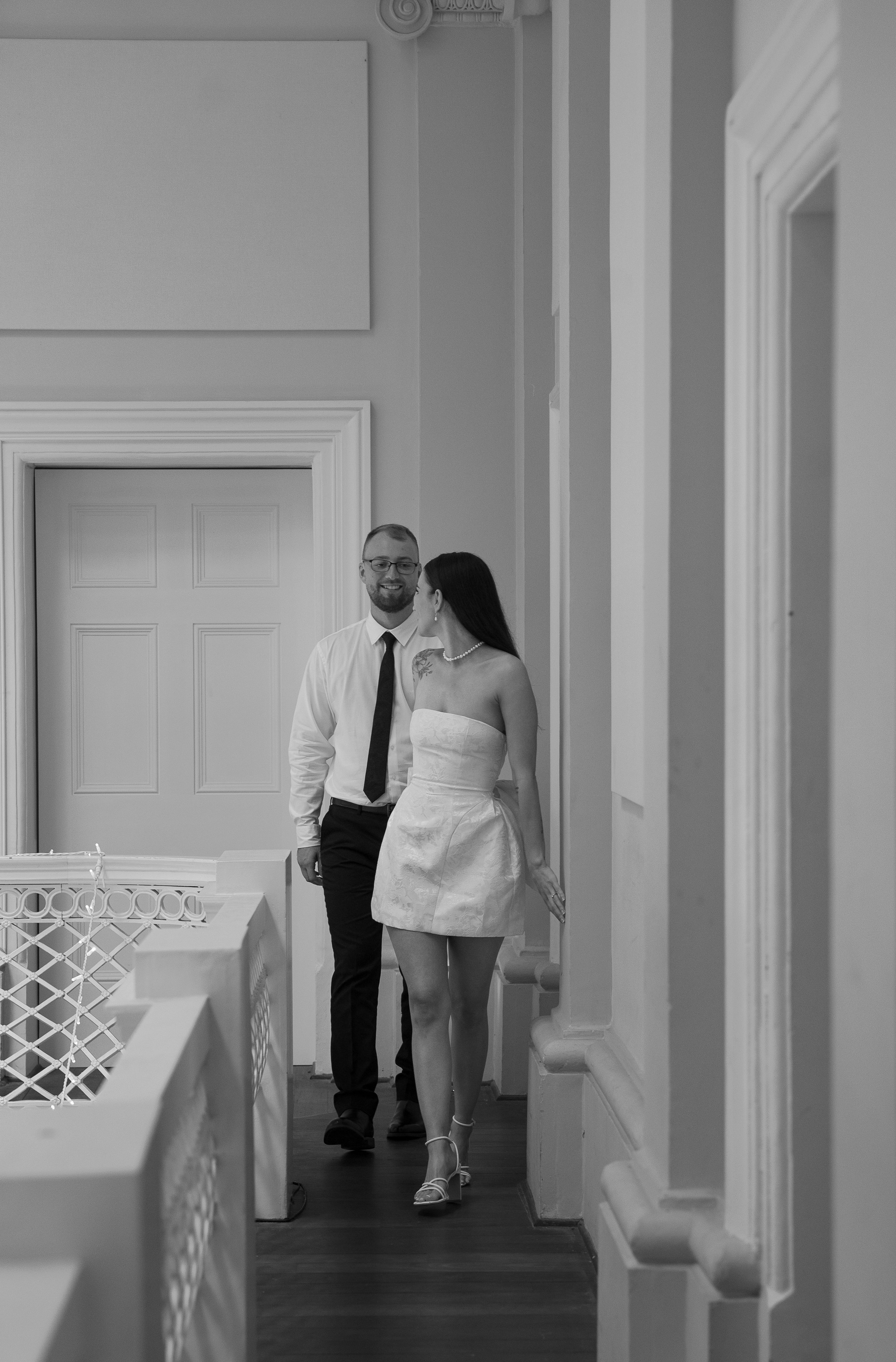 A man and a woman are walking down a hallway, smiling at each other. The woman is in a strapless white dress and high heels, the man in a white shirt, black pants, and tie. The scene is black and white.